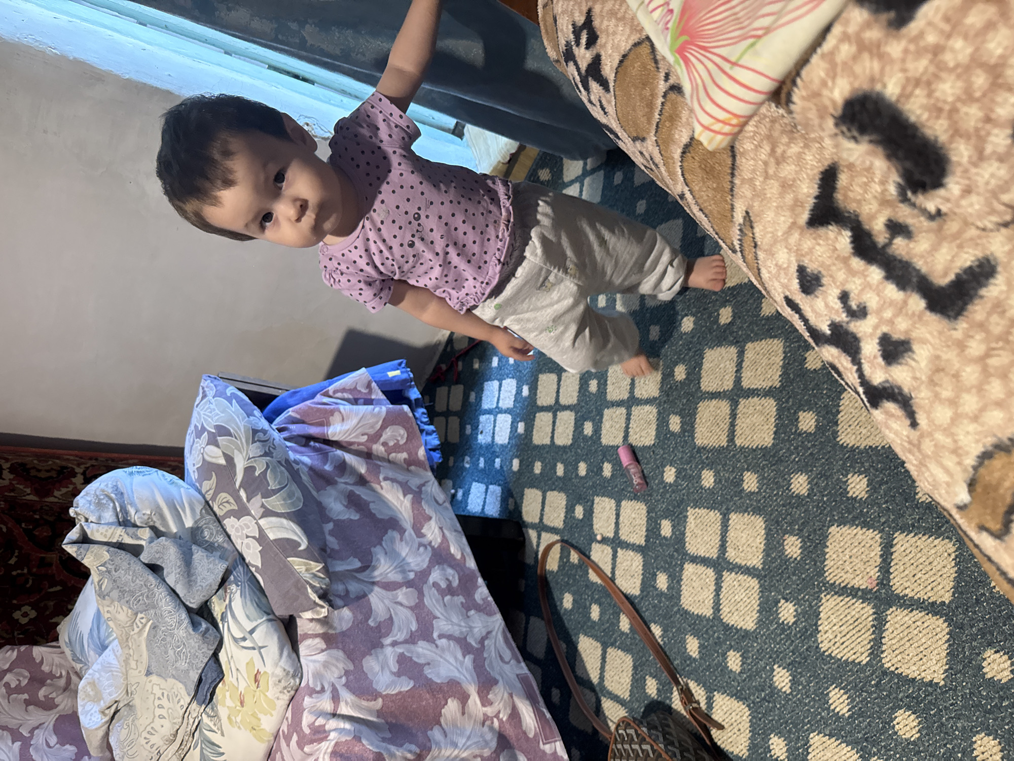 A toddler stands on a patterned rug in a bedroom in Chardara, Uzbekistan. The child, wearing a purple polka-dot shirt and gray pants, looks towards the camera with a neutral expression. To the left of the child, a bed is partially visible, covered with a purple and white floral comforter and scattered pillows. A brown handbag with a strap lies on the rug near the bed. A small pink tube, possibly a lipstick or balm, is also on the rug. The room appears dimly lit, with a window covered by a light blue curtain on the right side of the image. The walls are a neutral color.