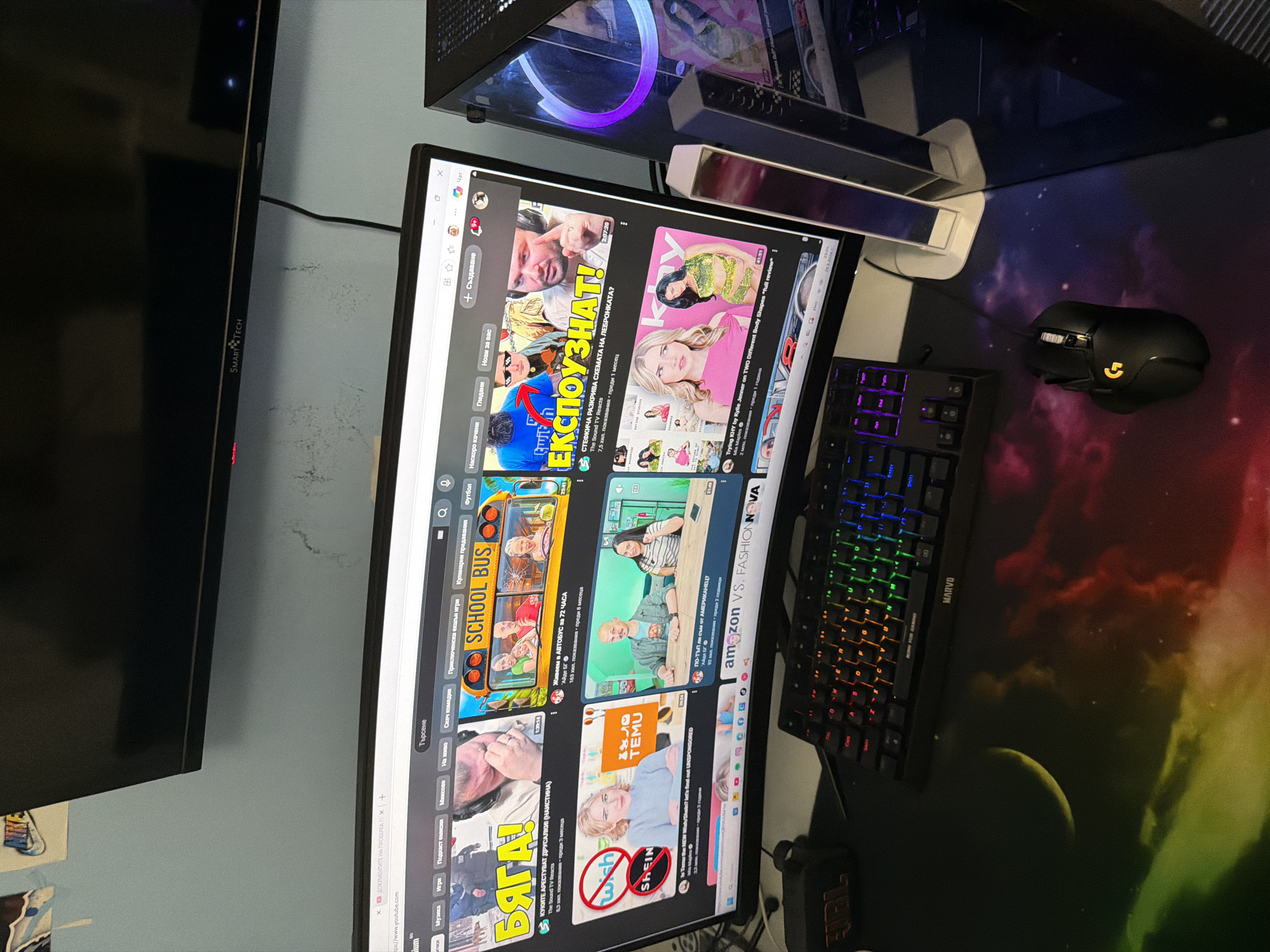 The image shows a desk setup with a computer monitor displaying the YouTube homepage. The monitor is positioned to the right of a black television. On the desk, to the right of the monitor, there's a gaming mouse, a keyboard, and a colorful mousepad featuring celestial imagery. In the top right corner, part of a computer tower with illuminated components is visible. The screen displays various YouTube videos in Bulgarian, including thumbnails featuring people and topics such as "school bus" and "Fashion Nova". The keyboard is a MARVO model with illuminated keys. The mouse is a Logitech model. The desk appears to be in a room with a wall painted in a light color, and the image is taken at a slight angle. It is impossible to determine the location in the city of Pernik, Bulgaria from this image.