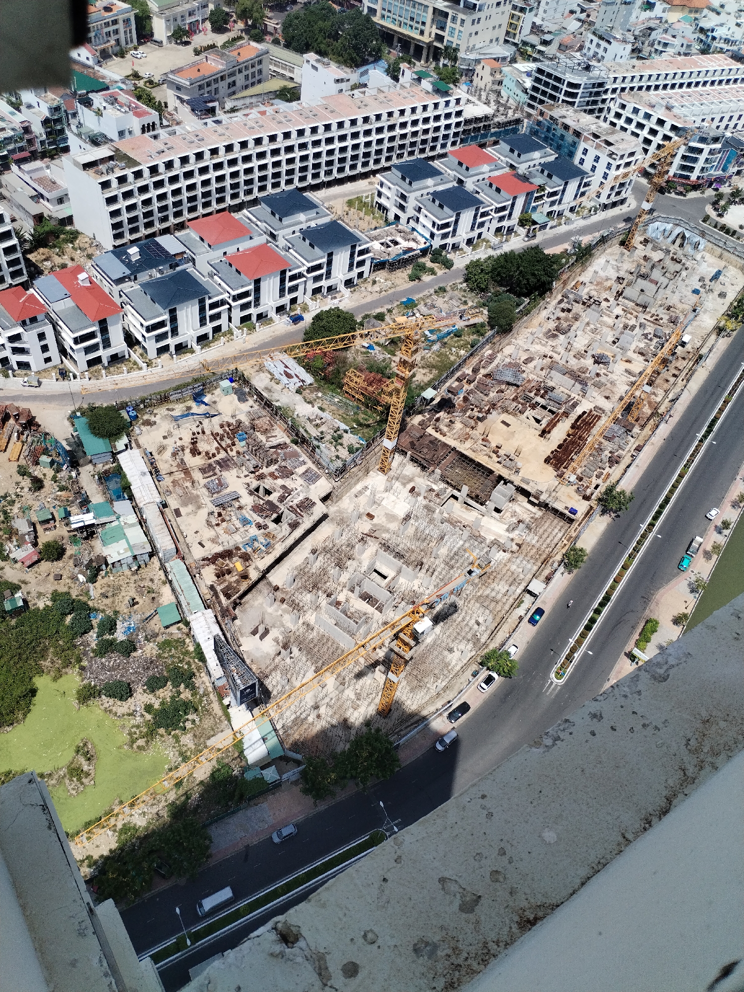 This is an aerial drone shot of a large construction site in a tropical urban environment, likely Nha Trang Ward, Vietnam. The scene is dominated by ongoing construction of multiple buildings. In the foreground, a partially constructed, large concrete structure is visible, with rebar and scaffolding indicating active work. Yellow construction cranes are prominent features of the site, one of them actively positioned over the concrete structure, suggesting ongoing lifting and placement operations.
Surrounding the central construction zone, there are newly built, modern residential buildings with white facades and dark or red roofs. These buildings appear completed or nearing completion, contrasting with the raw state of the main construction area. Some smaller, older structures with green corrugated roofs are clustered near the edge of the site, possibly temporary worker accommodations or existing structures that will be demolished.
A paved road runs alongside the construction site, with several vehicles, including cars and a truck, visible. The presence of trees and greenery, along with the bright sunlight casting shadows, indicates it is daytime, likely midday given the angle of the sun. The overall atmosphere is one of active development and urban expansion. The weather appears clear and sunny, typical of a tropical climate. No visible text is present in the image.