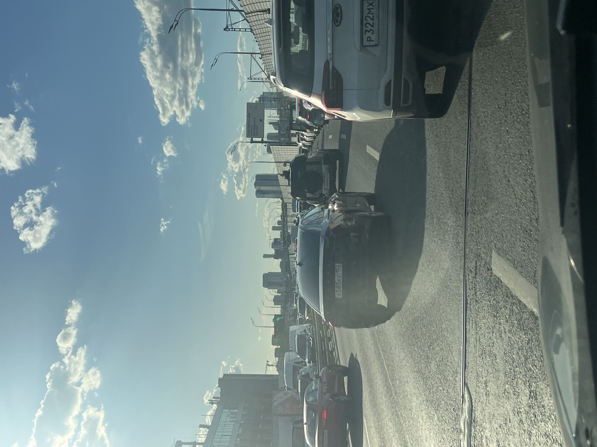 This media file depicts a heavy traffic jam on a highway in Moscow, Russia. The perspective is from inside a vehicle, looking forward and slightly to the side. The scene is dominated by a long line of cars and trucks on a multi-lane road. Several vehicles are clearly visible, including a white SUV with a license plate starting with "P322MX" and a black SUV with a license plate "X585KE152". Various other cars, vans, and trucks fill the road ahead, indicating significant congestion.
The time of day appears to be daytime, likely late morning or early afternoon, given the bright sunlight and the clear blue sky with scattered white clouds. The weather seems pleasant, with good visibility despite the traffic.
In the background, a cityscape with tall buildings is visible, typical of a major urban environment like Moscow. There are also infrastructure elements such as streetlights and highway signs. One sign in Russian appears to be an informational traffic sign, possibly indicating distances or directions to different areas.
The overall mood conveyed is one of frustration and patience due to the traffic. The camera is angled such that the road and vehicles are mostly seen from a slightly elevated perspective, looking down the highway. The sunlight casts shadows and highlights, adding to the depth of the scene.