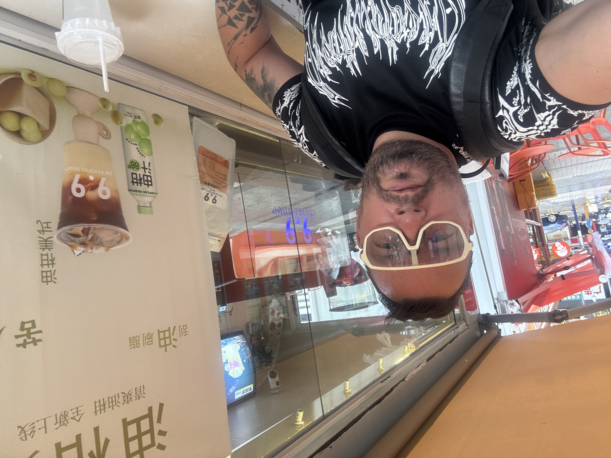 A man wearing white-rimmed sunglasses and a black t-shirt with white graphic designs is taking a selfie. He has dark hair and a beard. He is standing in front of a window of a coffee shop. The coffee shop appears to be located in Nanhai District, China, as indicated by Chinese text on a poster in the window. The poster displays images of drinks and fruit. Inside the coffee shop, there are tables and chairs, as well as other customers visible. Outside the coffee shop, there are red tables and chairs. A car is parked on the street in the background. A plastic cup with a straw is visible in the foreground.