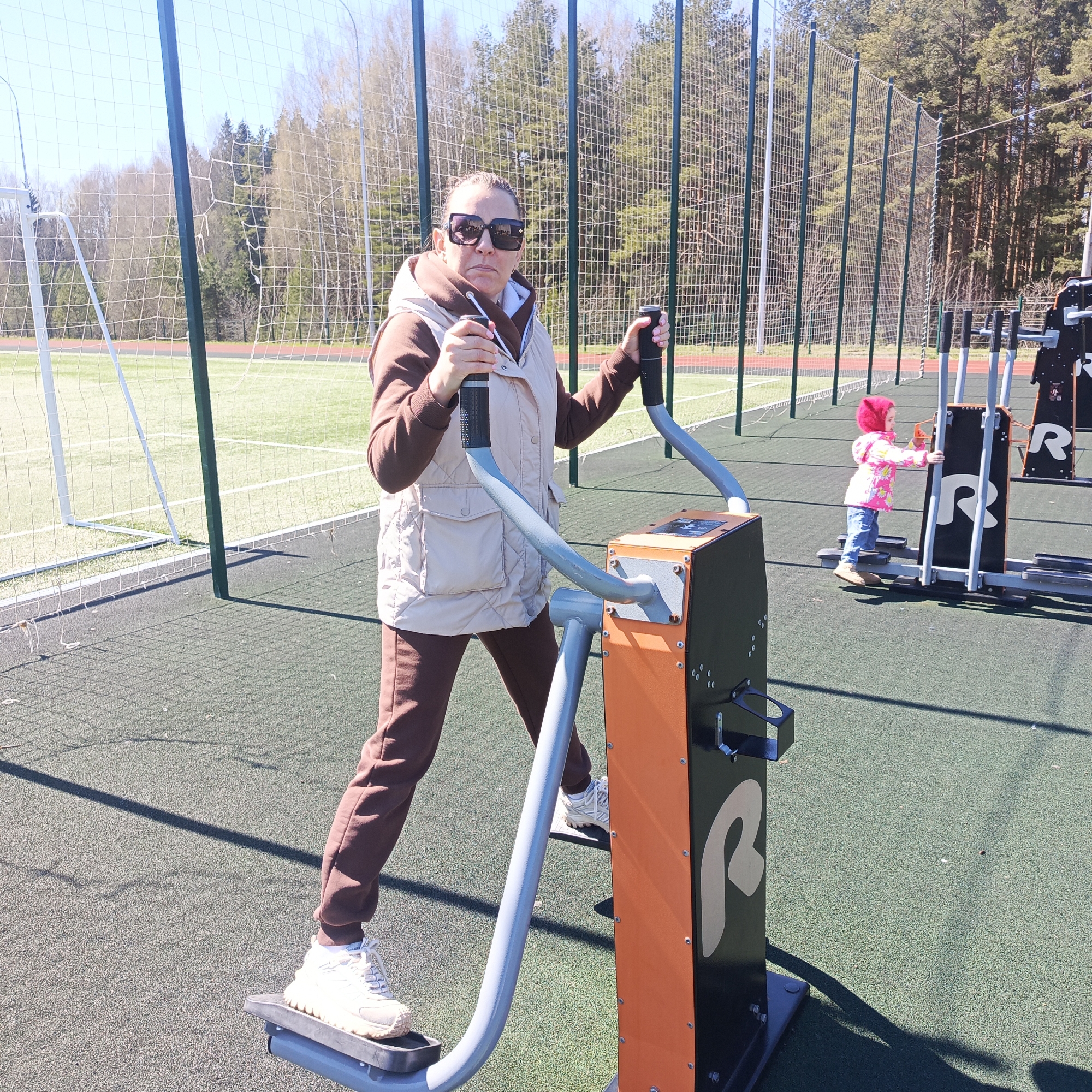 The image shows a woman using an outdoor exercise machine in Марковский, Russia. She is wearing sunglasses, a brown tracksuit, and a beige vest. To the right of the woman, a young child in a pink jacket and jeans is using a similar machine. Behind them, there is a sports field with a soccer goal and a fence. The background includes trees and a clear blue sky. The surface under the equipment is a green, rubberized material.