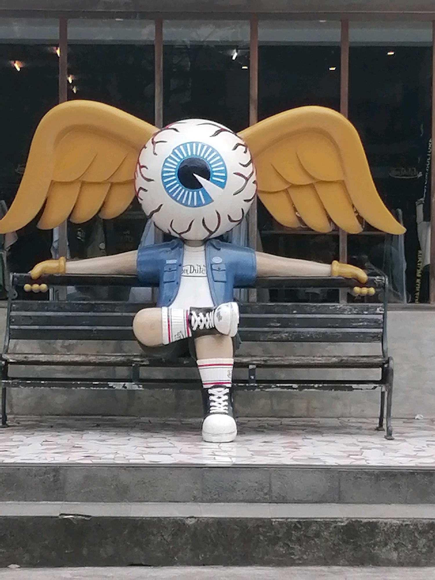 An outdoor shot captures a whimsical statue of a cartoon eyeball character with yellow wings, sitting on a park bench. The statue is dressed in a blue jacket over a white t-shirt that reads "Don Delle." It wears white socks with red stripes and black and white sneakers. The character's arms are outstretched, resting on the bench's armrests, and its legs are crossed. The background shows a building with large windows and a tiled pathway in front of the bench. The statue's playful and surreal design suggests it might be a public art installation in a creative urban setting. The area appears to be a public space, possibly a park or a street with seating.