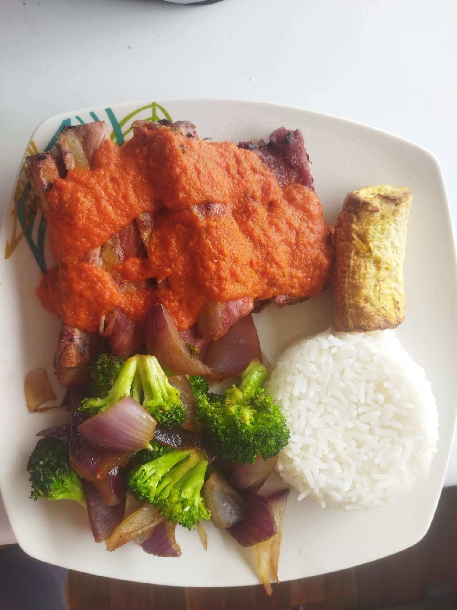 A plate of food consisting of ribs covered in red sauce, fried plantains, white rice, and sauteed broccoli and red onions is presented on a white, patterned plate. The location is Unknown, Unknown.