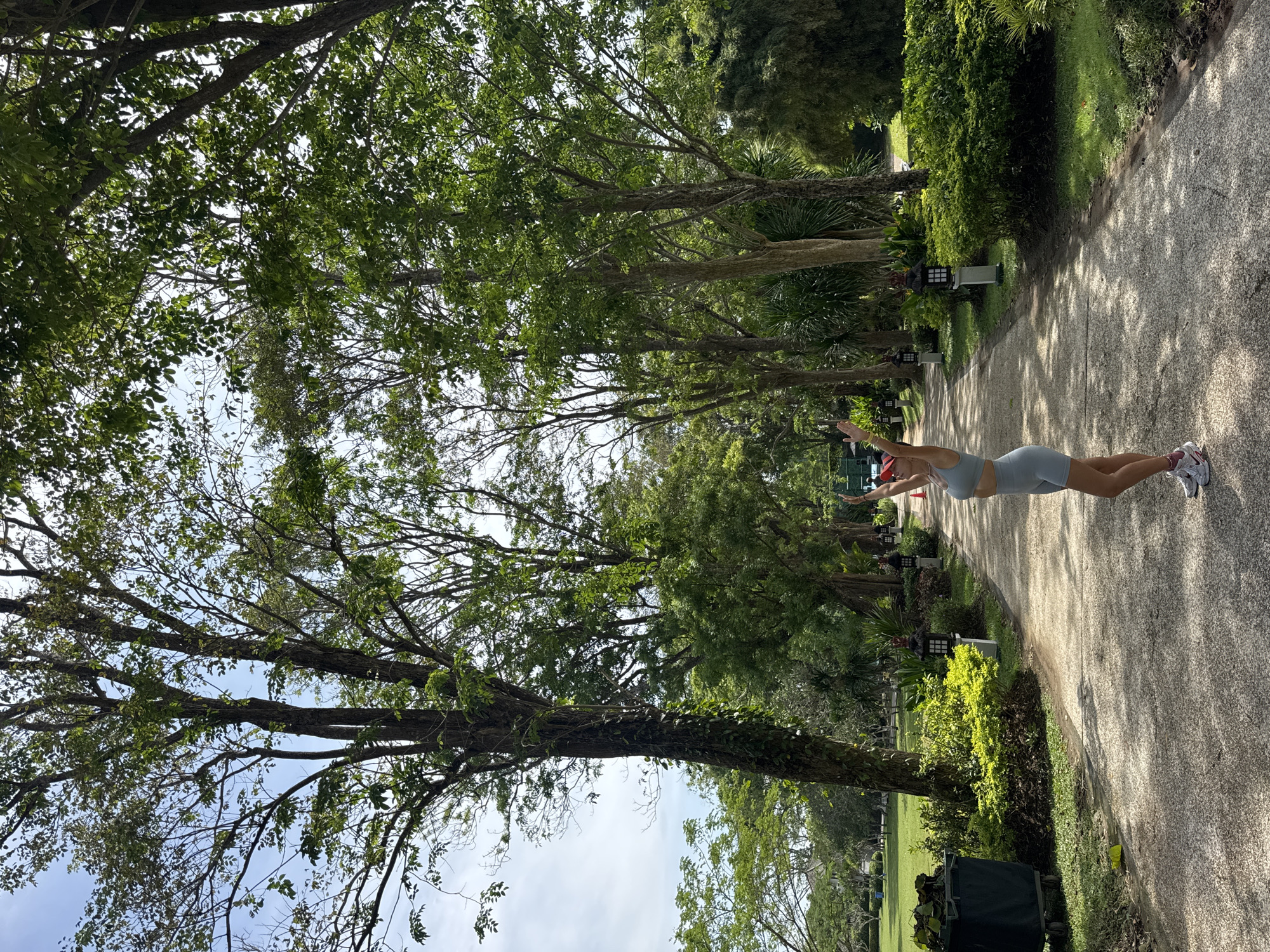 A horizontal, high-angle outdoor shot depicts a person in athletic wear on a paved pathway lined with trees and vegetation. The scene is located in Kuta Selatan, Indonesia.
The individual, seemingly female, is wearing a light blue athletic top and shorts set and athletic shoes. She stands toward the right side of the frame, arms raised outward, as if stretching or balancing.
The pathway, which runs diagonally from the lower right to the upper center of the image, is flanked by trees and lush green vegetation. Several ornamental Japanese lanterns are spaced along the path's edge. A dark green bin filled with leaves and garden debris is visible in the lower right corner.
The trees, with their dense green canopies, create a partial overhead cover for the path. Sunlight filters through the leaves, casting dappled shadows on the ground. The sky is partially visible through the branches, with patches of light blue.
