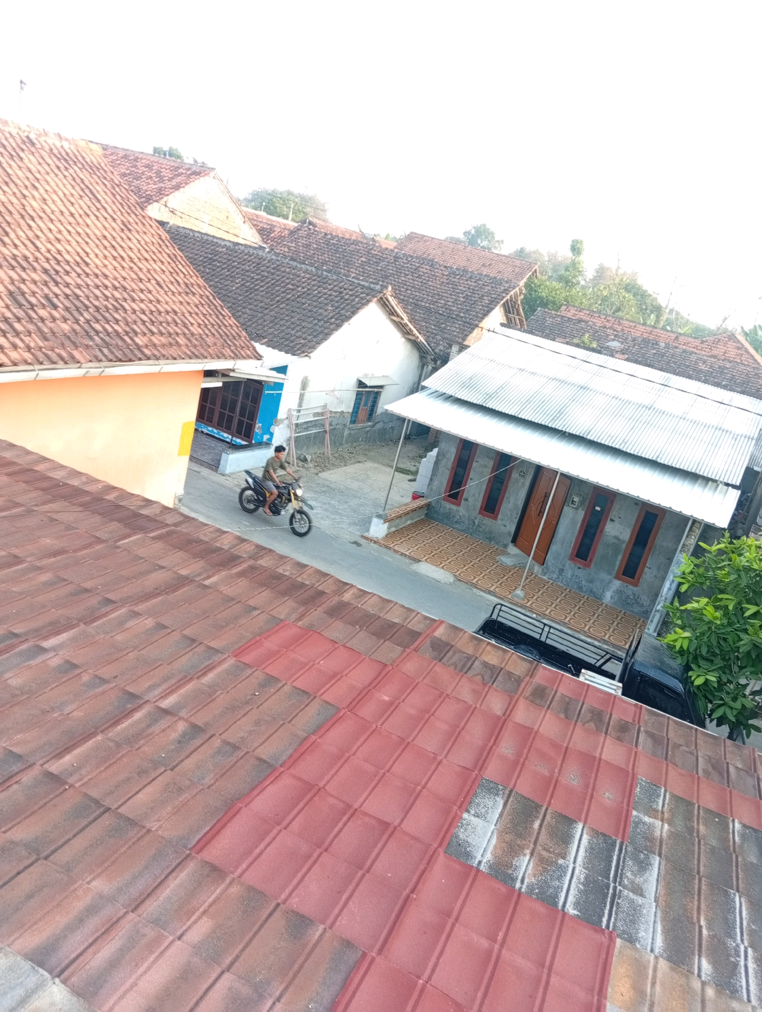 The image depicts a street scene in Kebandingan, Indonesia, viewed from an elevated perspective, likely a rooftop. In the foreground, a red tiled roof dominates the lower half of the frame. Beyond this, several houses with terracotta tiled roofs are visible, interspersed with modern structures featuring corrugated metal roofing. A man is riding a dirt bike along a paved street in front of a light-colored house. The street is lined with various houses, some appearing to be under construction or recently renovated. A tree with green foliage is visible to the right of the frame. The sky is bright and overcast, suggesting daylight conditions. The overall scene conveys a sense of a residential neighborhood in a tropical or subtropical climate.