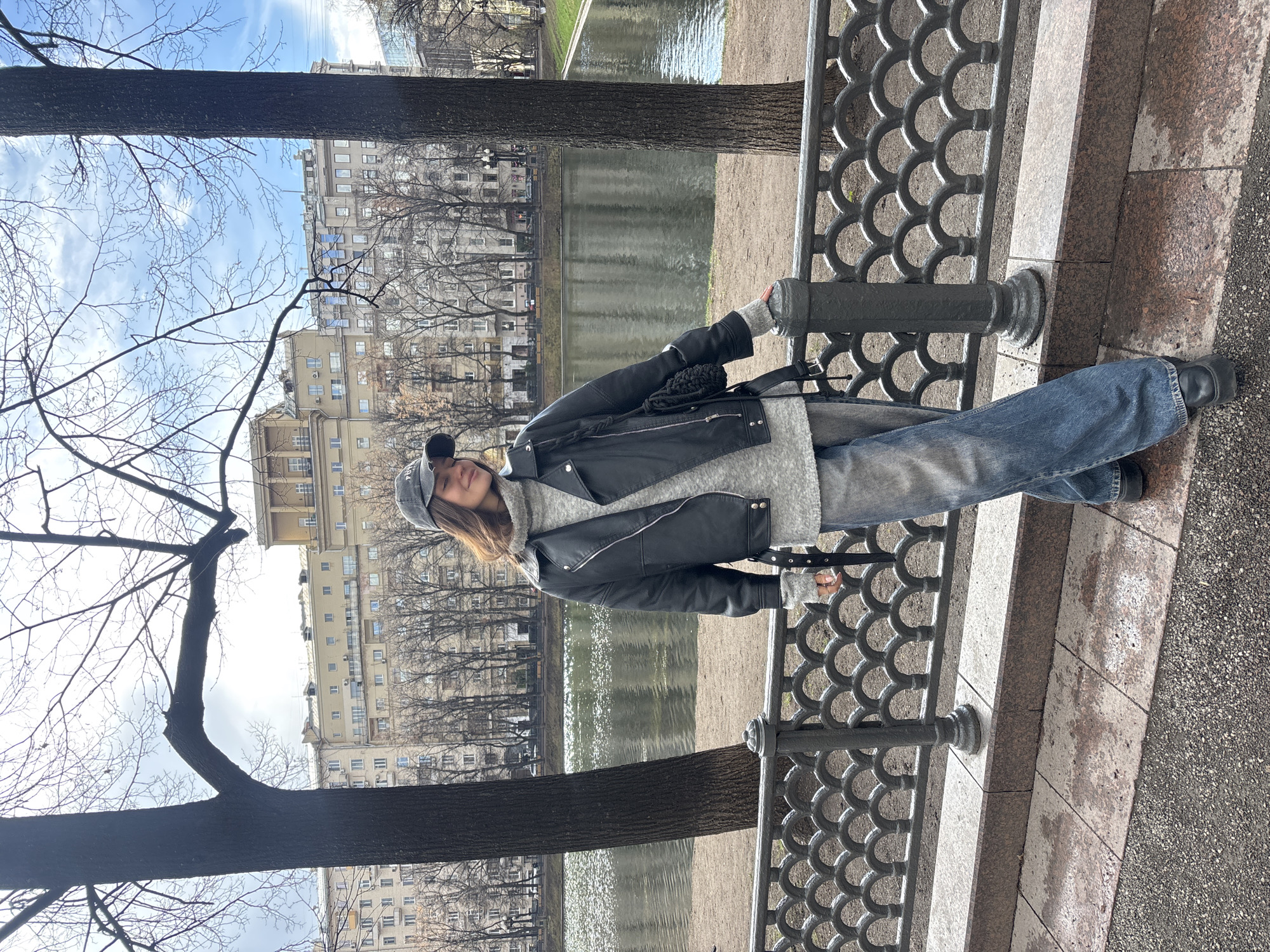 A young woman stands next to a decorative metal fence along a body of water in a public park. She wears a grey baseball cap, a black leather jacket, a grey fuzzy sweater, blue jeans, and black shoes. Her left arm rests on a metal pole, and she holds a small black purse. The park features leafless trees and a pond or canal with buildings visible in the background, suggesting an urban setting. Based on the architecture, this location is likely in Moscow, Russia.