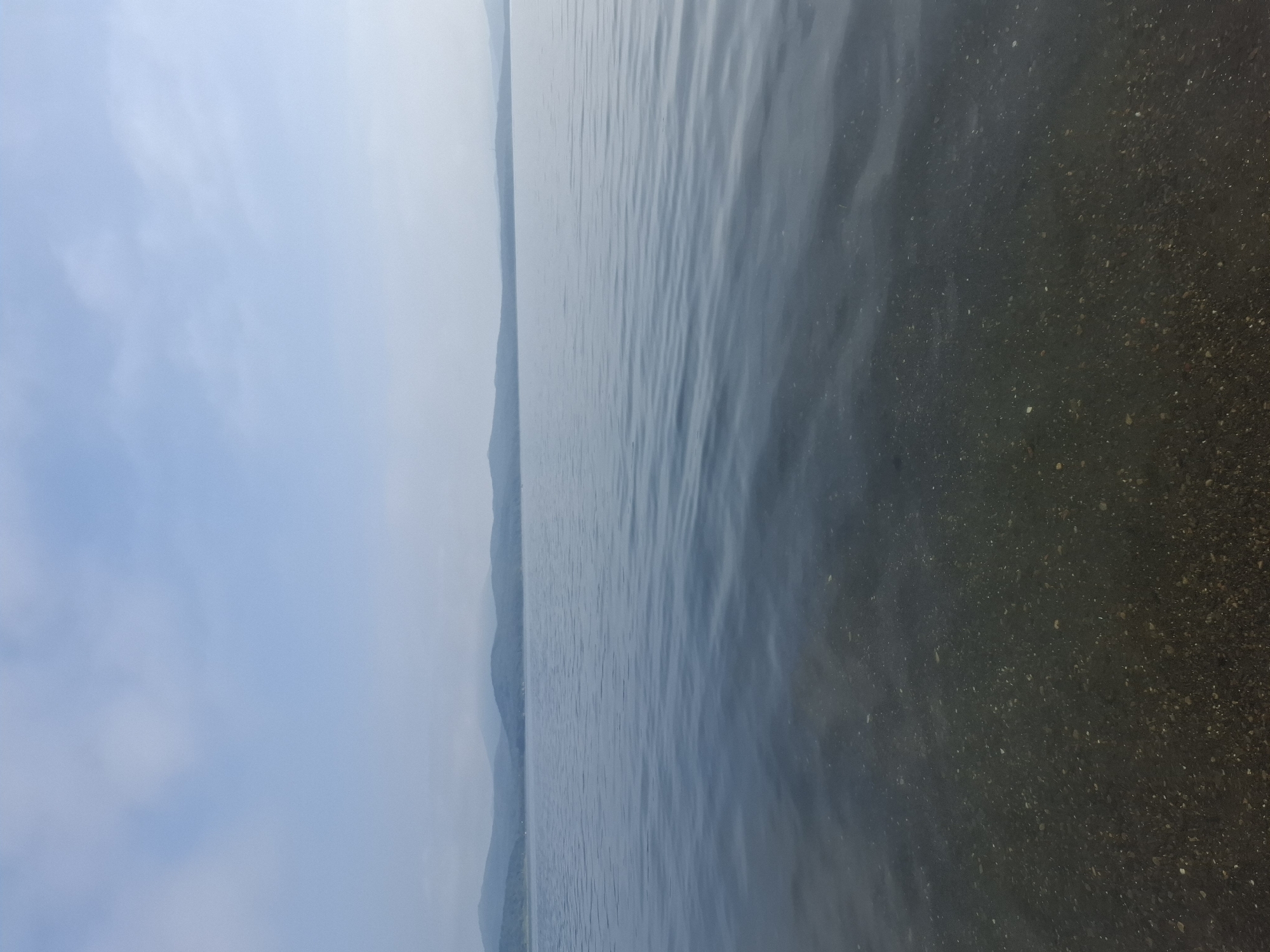 A serene landscape photo depicts a calm lake with rolling hills in the distance under a pale blue sky with scattered clouds. The foreground shows the clear, shallow water of the lake, revealing a pebbled bottom. The image conveys a sense of tranquility and natural beauty. There are no people or specific objects present. The location is not identifiable as Dunay, Russia, as there are no distinctive landmarks or features to indicate this specific place.