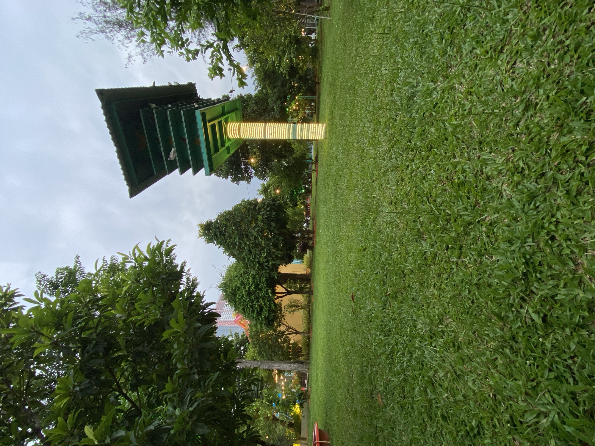 This image depicts a park scene in Nha Trang, Vietnam, at dusk. The foreground is dominated by a vast expanse of lush green grass, extending diagonally across the frame. To the left, a tall, multi-tiered green structure with a sloping roof stands prominently, resembling a pagoda or observation deck. Its supporting column is illuminated by a band of yellow lights, creating a striking visual contrast against the overcast sky.
Several white birds perch on the roof and ledges of the green structure, suggesting it might be a birdhouse or a feeding station. Behind the structure, various trees with dense foliage are visible, interspersed with small, decorative lights that add a festive ambiance to the park. In the middle ground, a vibrant, colorful building with geometric patterns can be seen through the trees, hinting at a larger urban landscape beyond the park.
The sky is a muted grey, indicating cloudy weather or the transition from day to night. The overall mood is peaceful and serene, with the illuminated structure and scattered lights offering a touch of warmth as daylight fades. There is no visible text in the image.