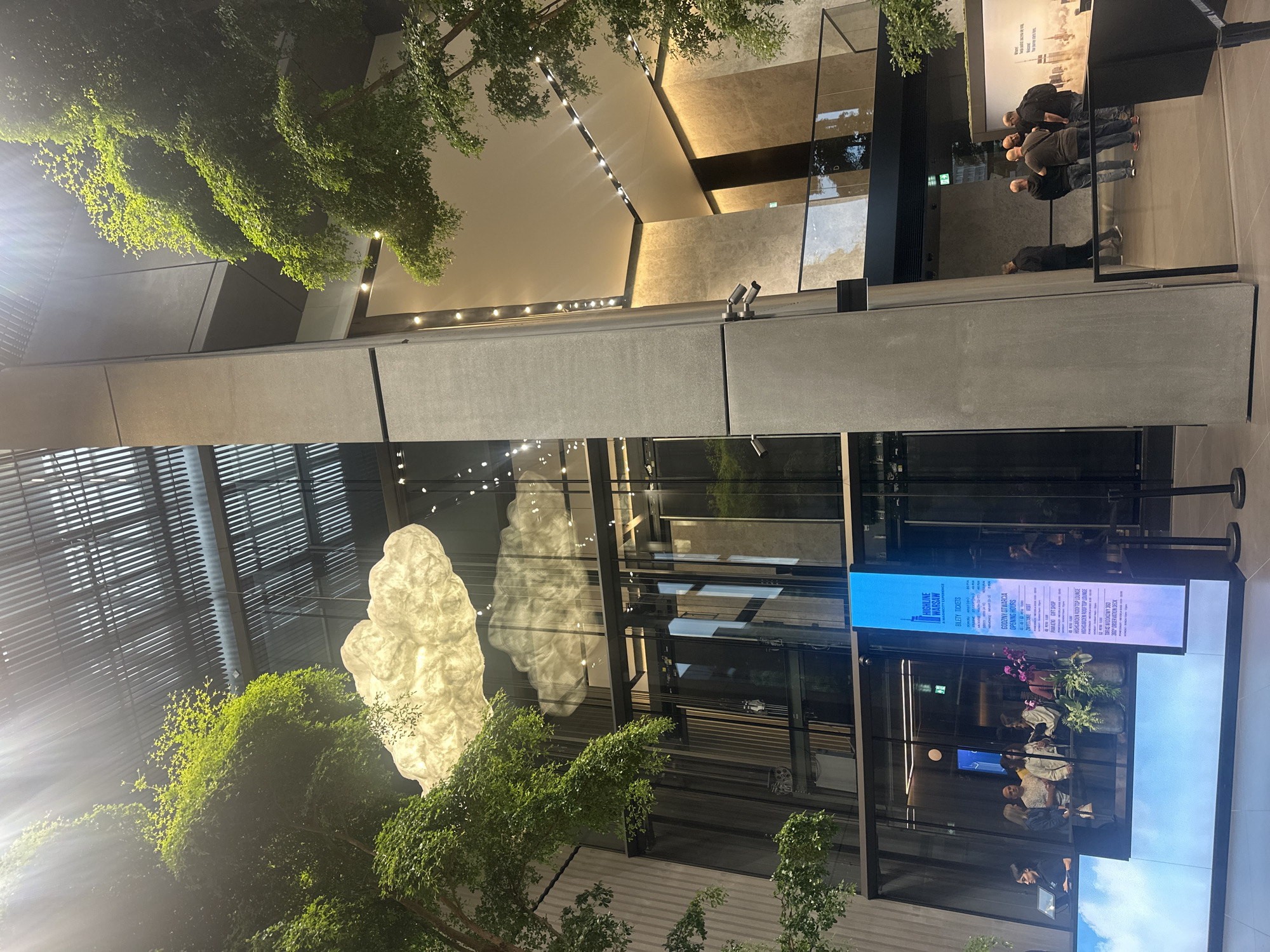 The image shows the interior of the High Five Warsaw observation deck, located in Warsaw, Poland. The scene is a modern atrium with a high ceiling and ample natural light filtering through a louvred roof. Large, artificial cloud sculptures are suspended from the ceiling, creating a whimsical ambiance. Two trees with lush green foliage are positioned on either side of the atrium, adding an element of nature to the urban setting.
In the foreground, a group of people are gathered near a ticket counter, suggesting they are either purchasing tickets or waiting for entry. A digital display board with information about ticket prices, opening hours, and attractions is prominently visible. The architectural style is contemporary, with concrete pillars, glass facades, and minimalist furniture. The reflection of the cloud sculptures in the glass elevator suggests the presence of a glass elevator within the atrium.
The overall impression is one of a sophisticated and inviting space, designed to offer visitors a unique and memorable experience of the city. The presence of greenery and the cloud-like installations contribute to a serene and airy atmosphere.