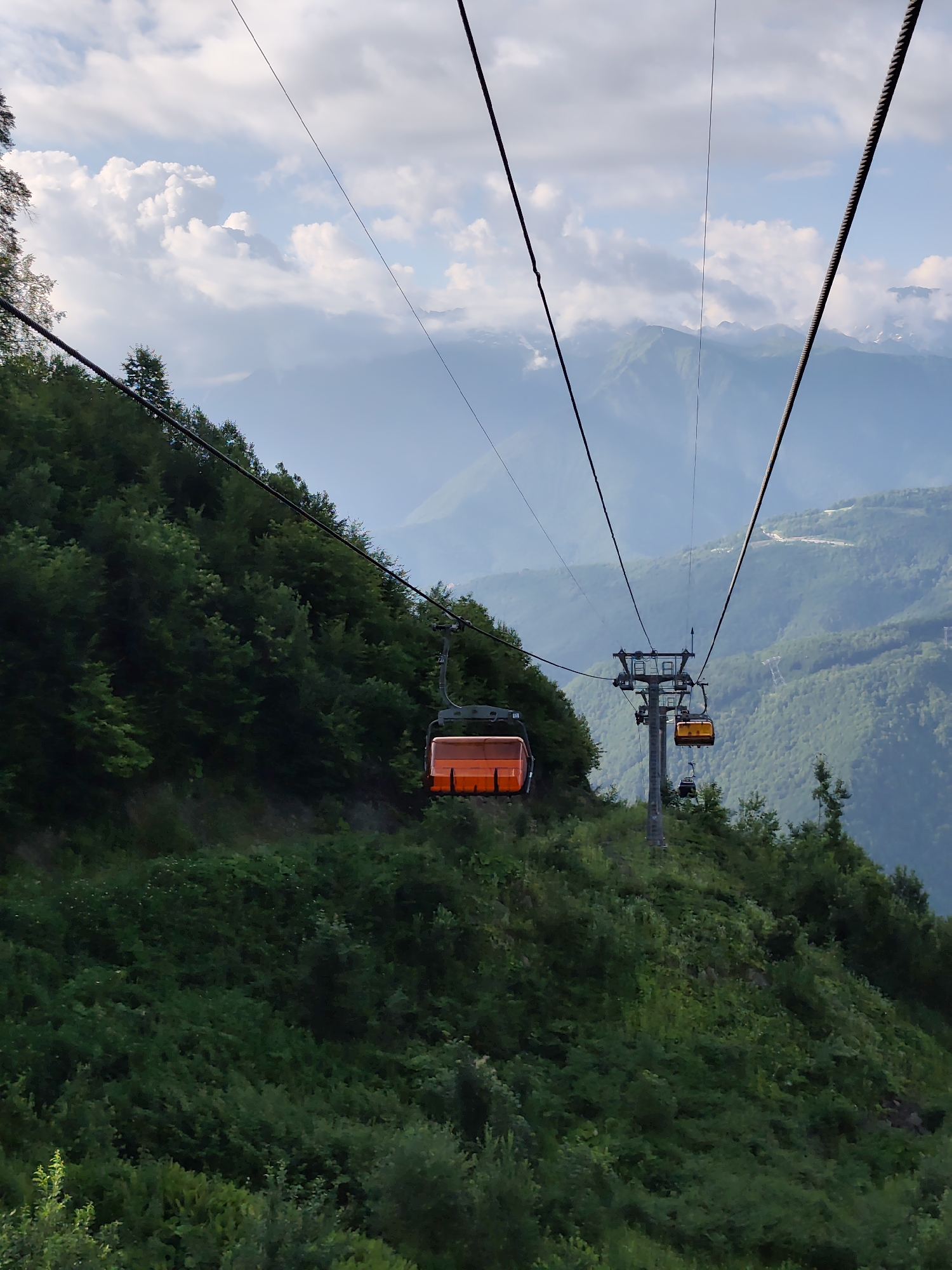 The image depicts a cable car ascending a lush, tree-covered mountainside in Estosadok, Russia. The cable car cabins are orange and yellow, suspended from thick cables that stretch towards the distant, misty mountain peaks under a cloudy sky. The foreground is dominated by dense green foliage, characteristic of a mountainous forest. The scene conveys a sense of travel and elevation amidst a natural, scenic landscape.