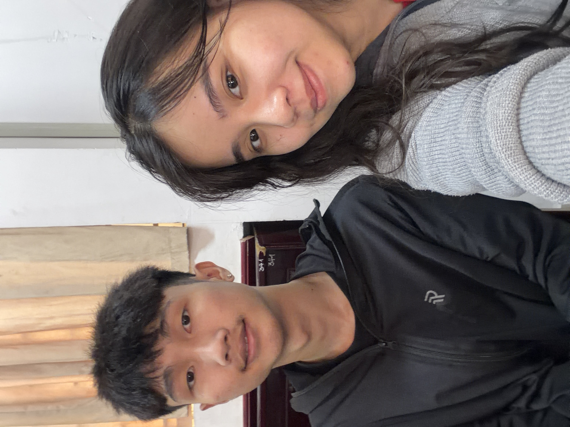Two individuals are visible. One is a young man, facing the camera, wearing a black zippered jacket. The second is a young woman, tilted towards the camera, wearing a grey sweater. The background appears to be an indoor setting with neutral-toned walls and wooden trim. The location is not identifiable as Đà Nẵng, Vietnam from the provided image.