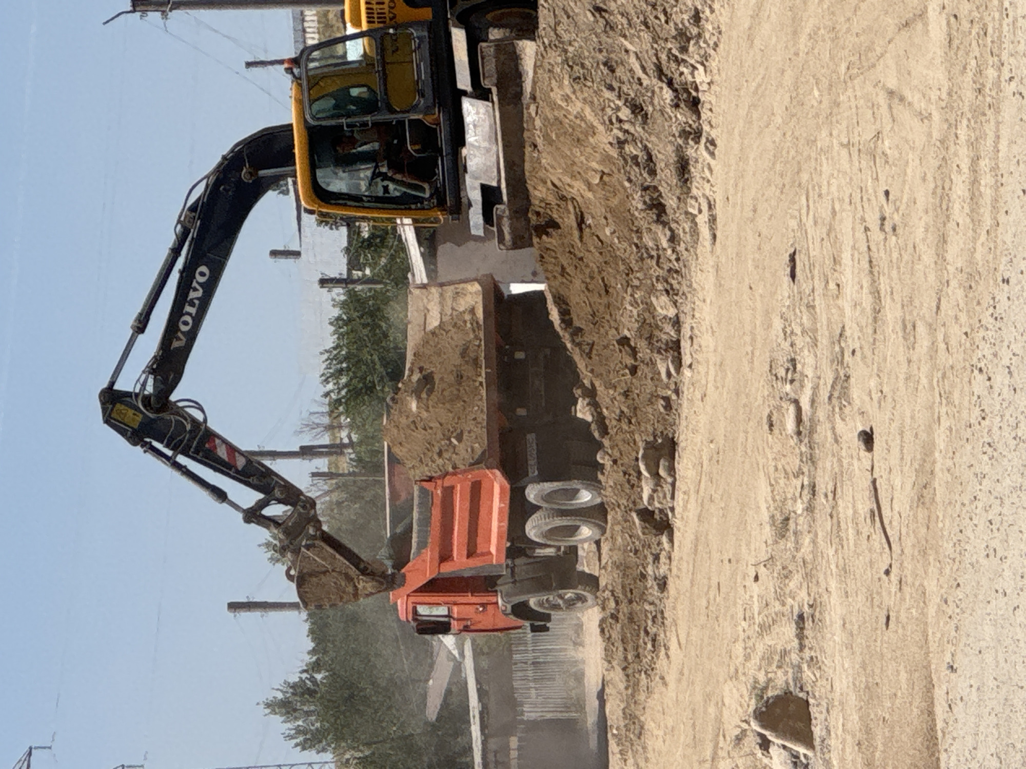 In Chirchik, Uzbekistan, an excavator is actively engaged in loading dirt into the bed of a red dump truck. The excavator, a yellow Volvo, is positioned to the right of the dump truck and its articulated arm is extended, scooping a load of dirt from a mound. The dump truck, facing left, has its bed partially filled with dirt. Both vehicles are situated on a dirt surface, indicative of a construction or excavation site. In the background, there are trees, utility poles, and some structures that appear to be part of a construction area. The sky is clear and blue.