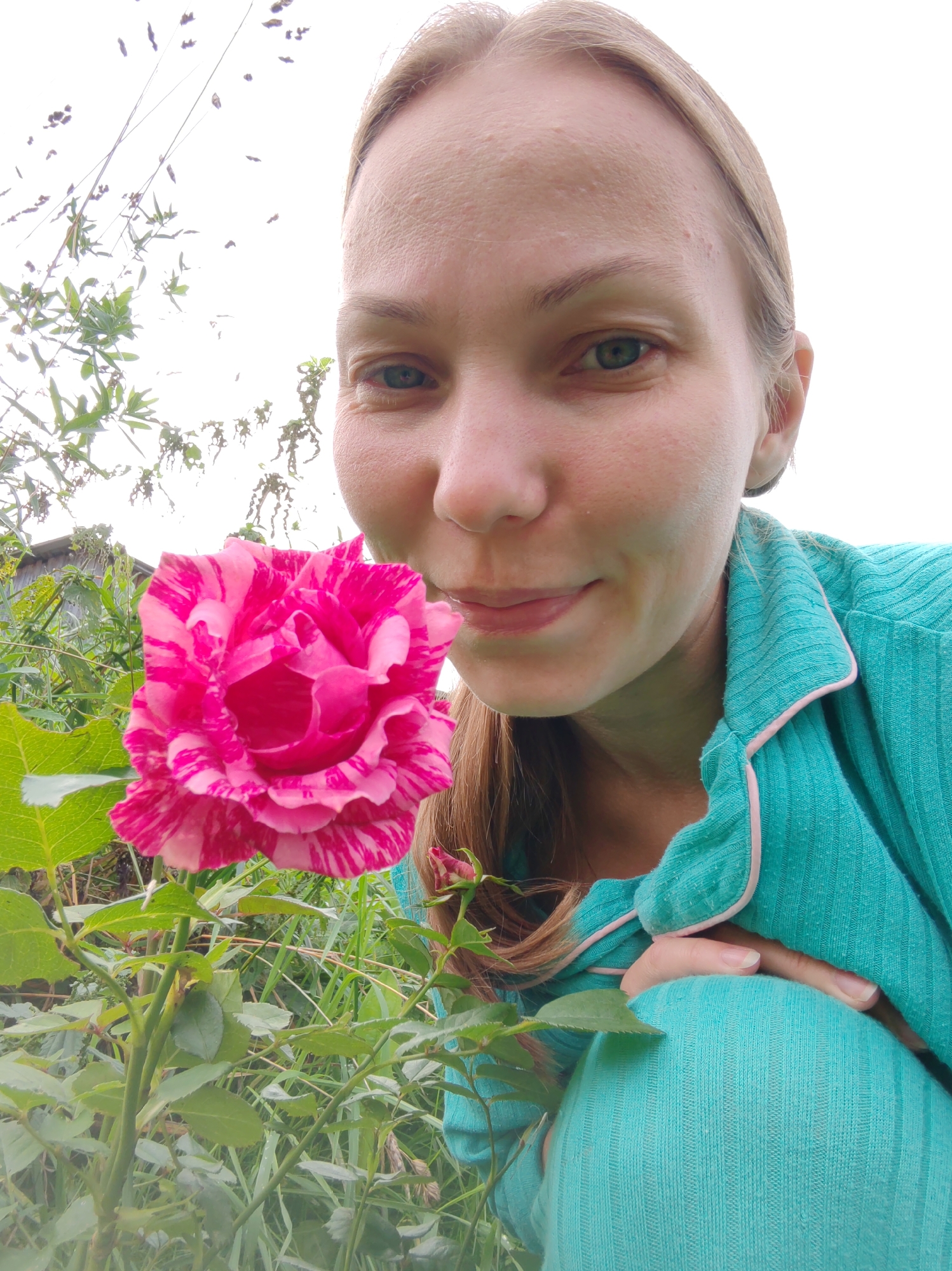 A low-angle, eye-level medium shot captures a Caucasian woman with fair skin and blonde hair, wearing a teal pajama set with pink trim, in an outdoor setting. She is leaning forward, bringing her face close to a vibrant pink and white striped rose. The woman's eyes are open, and she has a gentle smile, suggesting she is smelling the flower. Her right hand is visible, resting on her knee, which is also clad in the teal pajama material. The rose, in full bloom, is positioned in the foreground, to the left of the woman's face. Behind the rose and woman, lush green foliage and plants create a natural backdrop, with a hint of a wooden fence visible in the distance on the left. The sky above is bright and overcast. The scene appears to be in a garden or natural outdoor space, possibly a backyard. The location of Чухлэм, Russia, is not visually evident in the image.