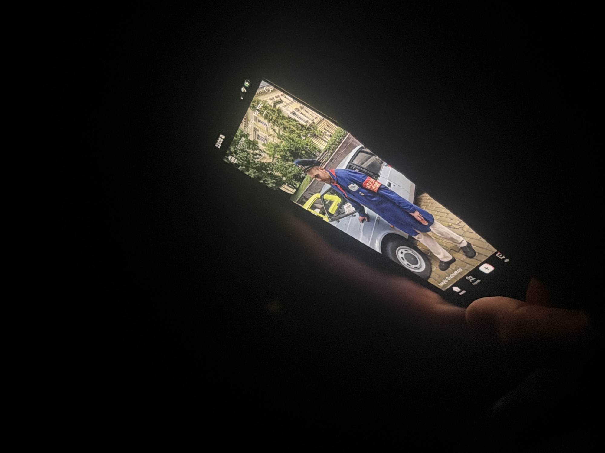 A photograph captures a person holding a smartphone in a dark environment. The phone's screen is brightly lit, showing an image of a man in a blue uniform standing beside a light-colored vehicle. The man, who appears to be a male, wears a blue cap, a blue jumpsuit or overalls, and has a prominent red armband on his left arm. He is also wearing light-colored trousers and dark shoes. Behind him, a multi-story building with numerous windows is visible, partially obscured by green foliage from trees.
The overall scene is dark, with the phone's display being the main source of light, suggesting a night-time setting. The top left corner of the phone screen displays "23:08," confirming the late hour. Below this time, the text "Explore. Following. For You" is visible, indicating a social media or content browsing application. At the bottom of the displayed image, the name "Ivan Gushlekov" is clearly readable. Further to the right, an icon resembling a heart or like symbol is accompanied by "8k," likely indicating 8,000 likes or interactions. Other interface elements like a plus sign and a share icon are also visible. A human hand, with fingers curled around the device, is partially seen on the right side of the phone, holding it.