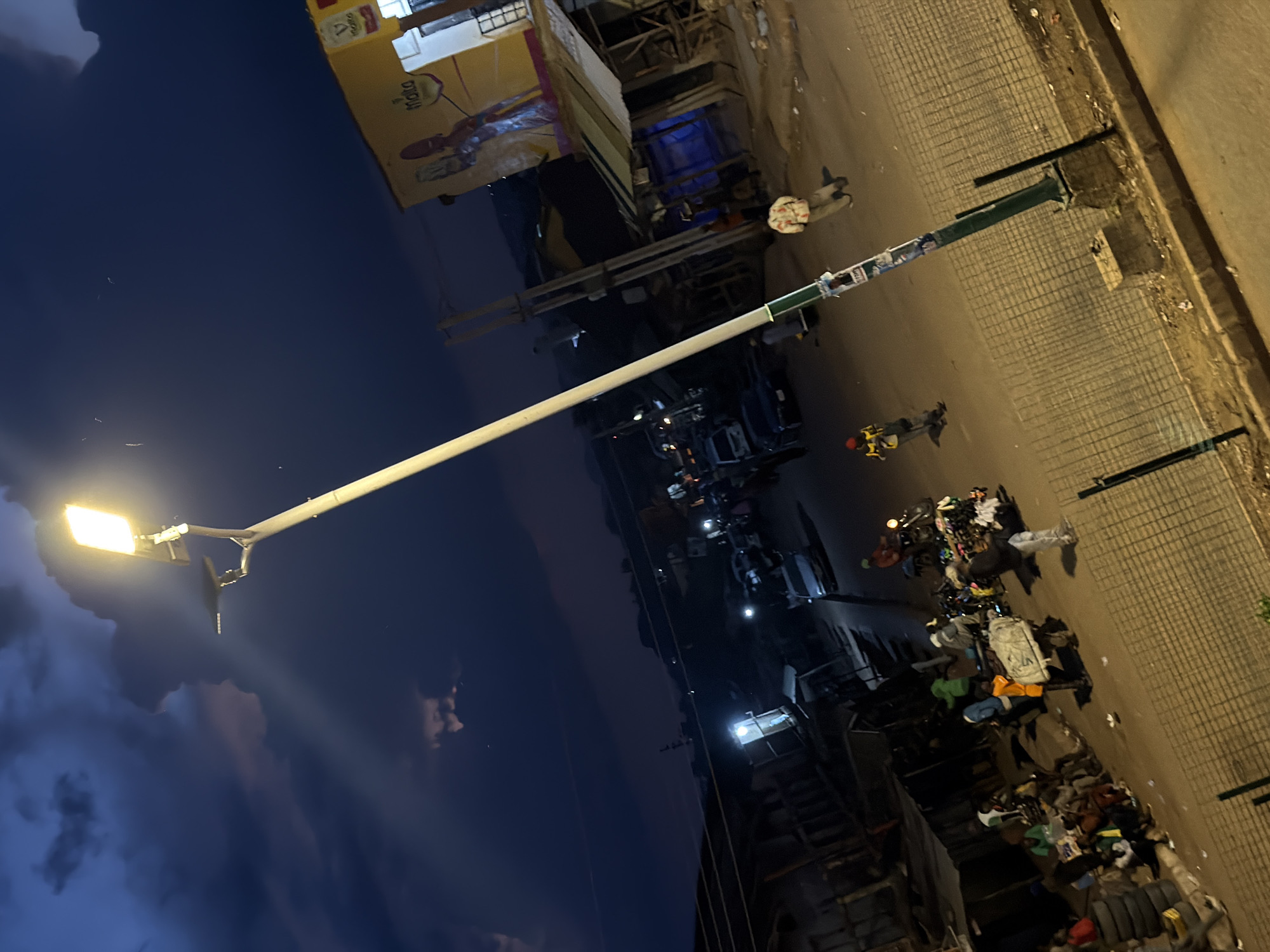 This is a high-angle, sideways shot of a street scene at dusk or night in Bukuru, Nigeria. The primary subject is a brightly lit streetlamp extending from the bottom left to the upper right of the frame. The lamp illuminates a section of a paved road with a patterned texture, likely cobblestone or interlocking pavers.
On the right side of the road, there is a building with a yellow awning that has text that is difficult to read but appears to be in a dark color. Below the awning, the building has a darker facade. Adjacent to the building, a narrow walkway or sidewalk is visible, lined with what appears to be trash and various objects.
The street itself shows evidence of activity. Several people are visible, some walking and others near parked motorcycles or carts laden with goods. The objects on display on the roadside include bags, clothing, and what looks like a display of items for sale. The overall impression is of a busy market or street vending area in a Nigerian town.
The sky is a deep blue, indicating either late evening or early morning. There are some clouds visible in the sky, some of which are illuminated by the streetlamp's glow, creating a dramatic effect. The lighting suggests it's a time when artificial lights are needed, amplifying the contrast between illuminated areas and shadows. There are no clear indicators of weather conditions other than the presence of clouds. The scene conveys a sense of everyday life and commerce taking place under artificial illumination.