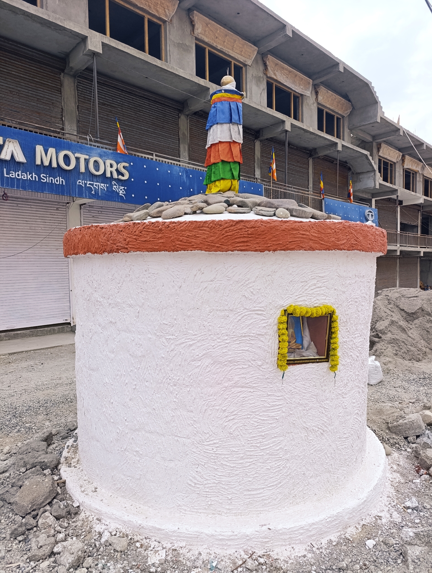 In Leh, India, a white cylindrical structure with a textured finish and a reddish-orange rim stands prominently in the foreground. The structure is adorned with a small framed image, surrounded by yellow flowers. The top of the structure is covered with small, rounded stones and topped with a colorful stupa-like ornament featuring layers of blue, white, red, orange, and green fabric, topped with a golden finial. Behind the structure, a building under construction is visible, with unfinished concrete floors and walls, and roller shutters covering the ground-level entrances. A blue signboard with the white text "TATA MOTORS" and "Ladakh Sindh" in English and Tibetan script is mounted on the building. Prayer flags in orange, white, and blue are also visible, attached to the building. The ground around the structure is uneven and covered with gravel, broken concrete, and debris, suggesting a construction site. In the background, a large pile of sand or gravel is visible. The sky is overcast.