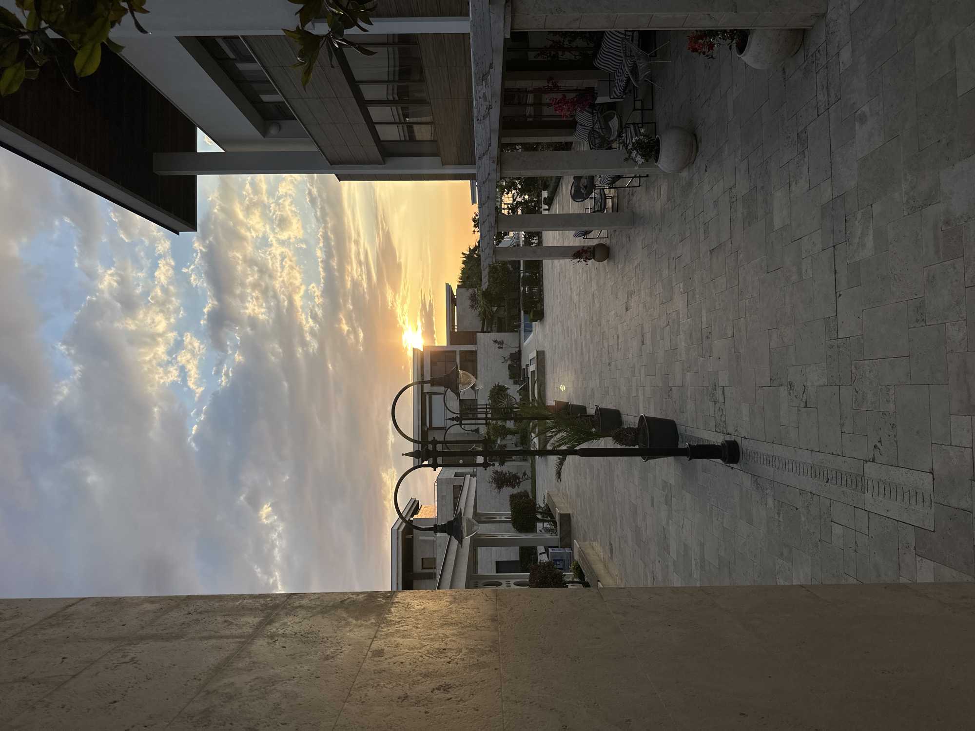 This is a photograph taken at sunset in Sozopol, Bulgaria, likely from a courtyard or pathway of a modern residential complex or hotel. The setting sun casts a warm glow across the sky, illuminating scattered clouds with hues of orange, yellow, and soft blue. The foreground is dominated by a paved courtyard made of light-colored, irregular stone tiles. To the left, a large, textured stone wall forms a significant portion of the frame.
A row of black, ornate lampposts with clear glass lanterns lines the pathway, each accompanied by a potted plant, some appearing to be palms. In the mid-ground, several modern buildings with clean lines and large windows are visible. One building on the right features a dark, slanted roof overhang supported by white columns. A covered outdoor seating area with striped chairs and tables is set up under a pergola-like structure. Potted plants with red and white flowers are placed around this seating area.
Further back, more buildings and landscaping are visible, including what appears to be a swimming pool. The distant horizon shows a sliver of the sea, with more buildings and greenery. The overall atmosphere is one of calm and luxury, characteristic of a resort setting during the golden hour. There are no people visible in the image.