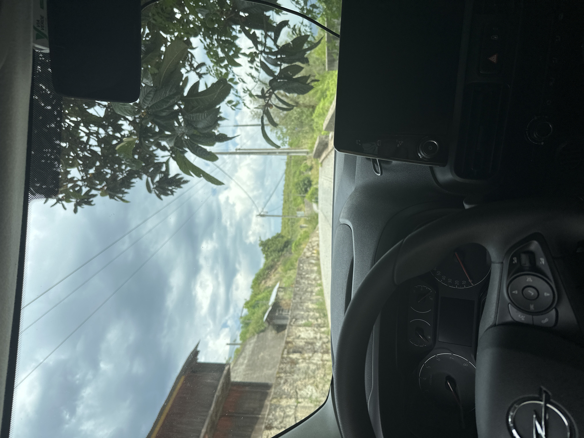 The image is taken from inside a car, with the dashboard and steering wheel prominently visible. Outside the car window, a street scene unfolds in Amarante, Portugal. A cloudy sky hangs above a road lined with vegetation. There's a structure that appears to be a stone wall or building along the roadside. The viewpoint suggests the car is either parked or moving slowly on a road in a somewhat hilly or elevated area of Amarante. The focus is divided between the car's interior and the landscape visible through the windshield.