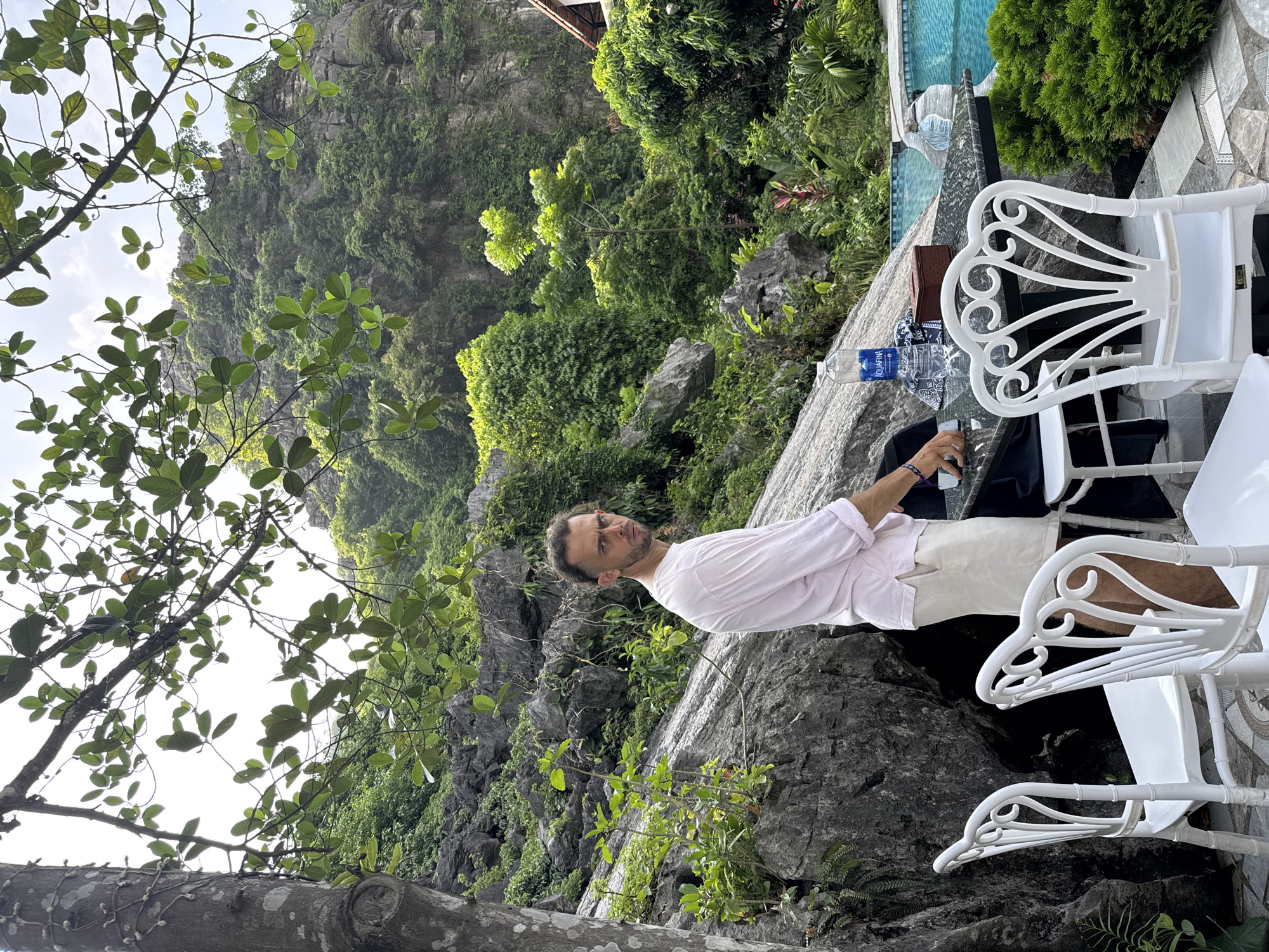 In the city of Nam Hoa Lư Ward, Vietnam, a man stands by a rocky cliffside with lush vegetation. He is wearing a light pink long-sleeved shirt and light-colored shorts. In front of him, a table is set with white chairs, a bottle of Aquafina water, a small brown box, and a cell phone. The scene is complemented by the natural beauty of the surroundings, with a swimming pool visible in the background.