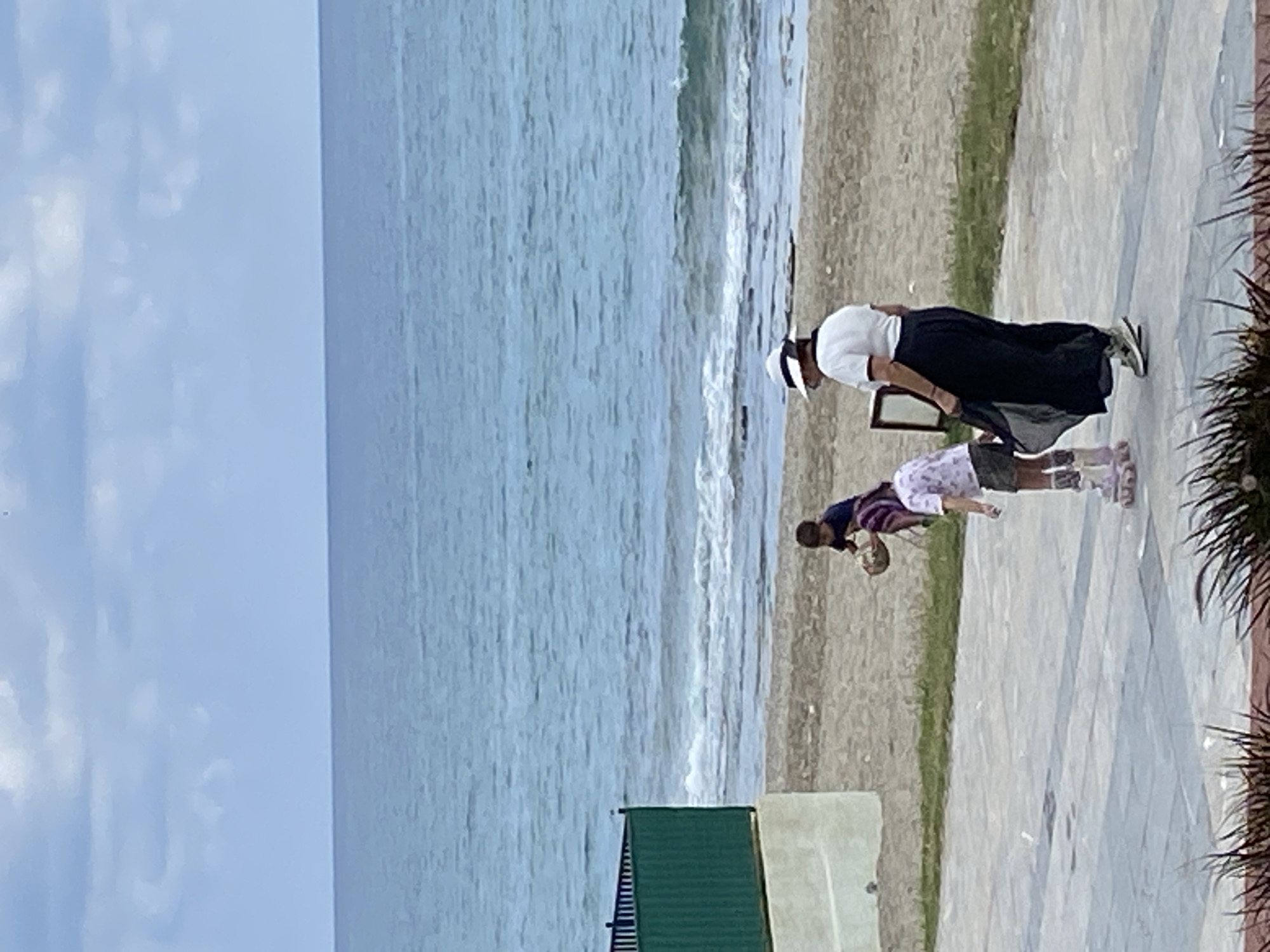The image shows a seaside scene in Alanya, Turkey. The view is from a slightly elevated position, overlooking a beach with the ocean stretching out to the left. A person wearing a white shirt, dark skirt, and a hat is bending over, possibly picking something up. A child is standing beside them, also looking down. They are on a paved area with some greenery in between. The waves are visible along the shore, and the sky above is partly cloudy. In the lower part of the image, there's a building with a green roof near a beach hut.