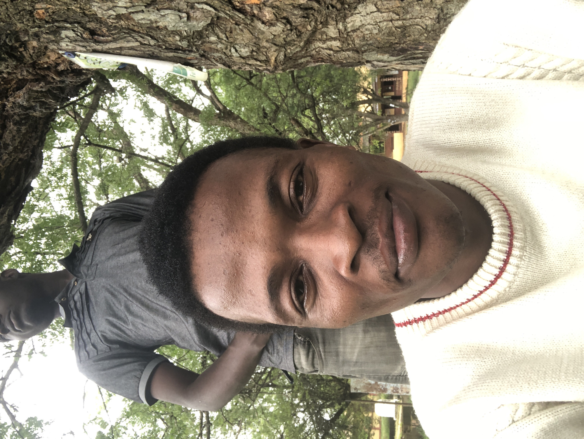 A low-angle, eye-level shot captures two Black men in an outdoor setting, partially obscured by a tree. In the foreground, a man in a cream-colored cable-knit sweater with red trim smiles directly at the camera. His face is centered, with his eyes, nose, and mouth visible. Behind him, the upper torso and head of another man are visible, looking upwards and away from the camera. He wears a dark grey polo shirt. The background shows a textured tree trunk on the right, with its bark prominently featured. Beyond the tree and the men, lush green foliage from trees and branches creates a dappled canopy, suggesting a park or wooded area. In the further background, a building with a red-tiled roof is visible through the trees, indicating a suburban or semi-rural environment. There is a small, white object with some writing on it attached to the tree trunk.