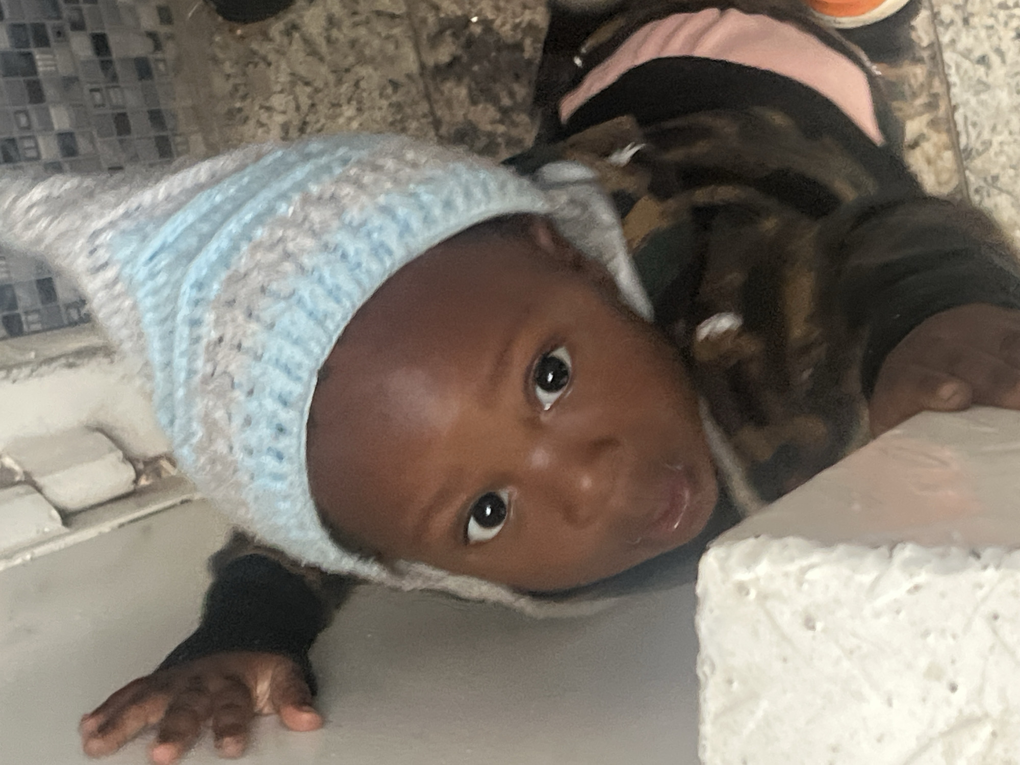 A baby wearing a blue and grey knit cap lies on a light-colored surface with a white brick in the foreground. The baby's head is tilted, and they are looking up at the camera. The baby's skin is brown. A small hand is extended to the left. The surface appears to be tiled in grey and white. It is not possible to determine if the image was taken in Gura Top, Nigeria.