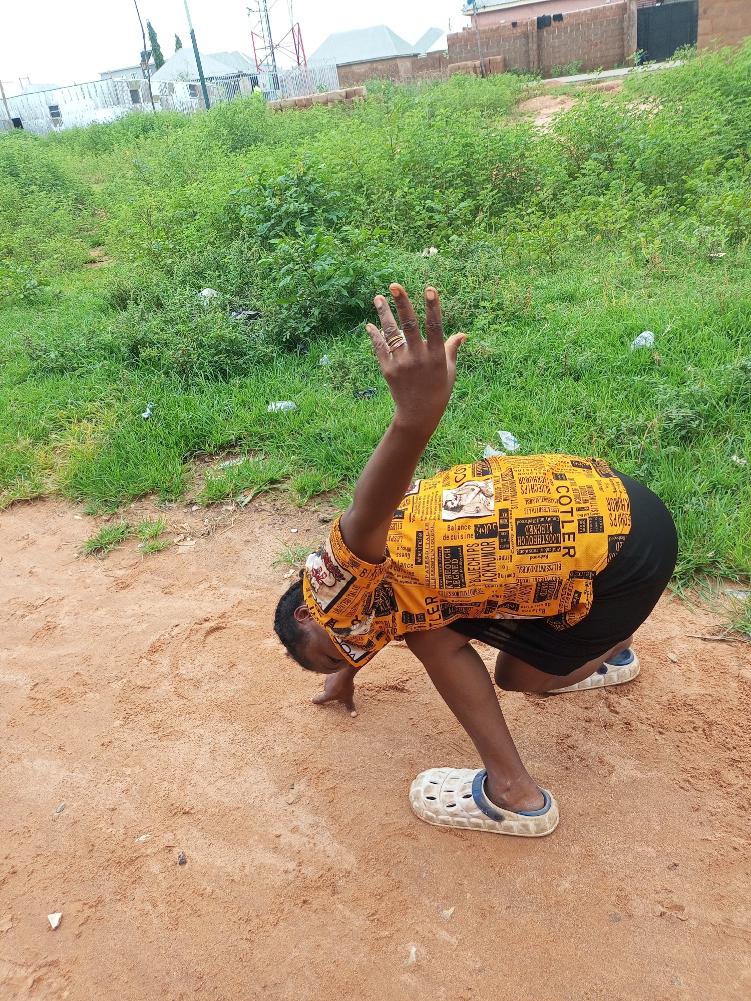 A person is crouched low on sandy ground, facing away from the camera, with their right arm raised upwards and their left hand on the ground for support. They are wearing a bright yellow shirt with a black graphic print and a black skirt. The person is also wearing white clogs with blue trim. The background consists of overgrown green grass and foliage, with some buildings and structures visible in the distance. The sky is overcast. The ground is composed of reddish-brown sand with a few small pebbles and debris.