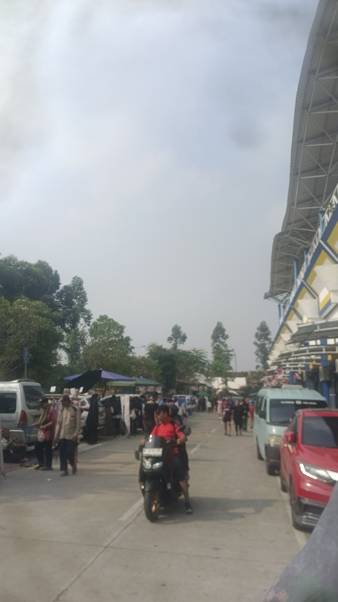 This is a vertical, medium-distance, eye-level shot of an outdoor market scene on a cloudy day in Tangerang, Indonesia. In the foreground, a person wearing a red shirt is riding a black motorcycle. Behind them, several other people are walking and standing. To the left, a white van is parked, and further back, there are market stalls with people browsing. To the right, a light blue van and a red car are parked on the side of the road. The background features a large, modern structure with a grey and yellow roof, possibly a stadium or convention center, and lush green trees. The overall scene depicts a busy marketplace with vehicles and pedestrians.