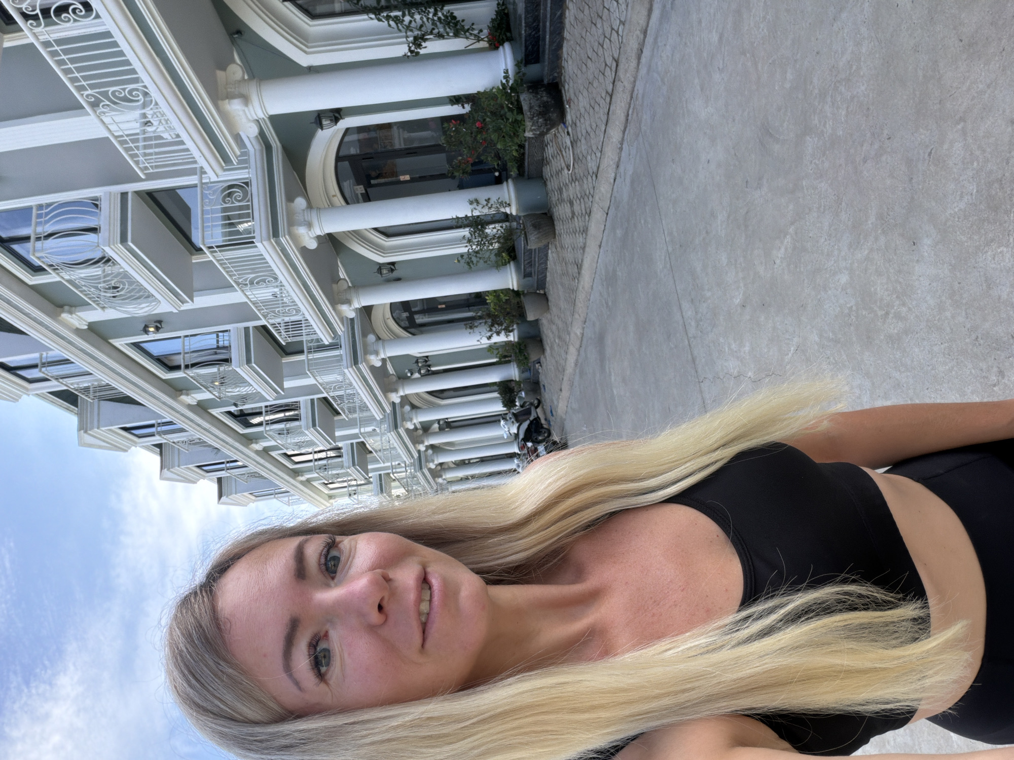 In a low-angle, selfie-style photograph taken outdoors on a paved walkway in front of a building, a blonde woman is seen from the chest up, looking towards the camera. She has light skin, blue eyes, and her long blonde hair is partially obscuring her face and shoulders. She is wearing a black sports bra and what appears to be black workout leggings.
The background features a multi-story building with a light blue facade and white decorative trim. The building has numerous balconies with white ornate railings and large arched doorways on the ground floor, framed by white columns. Potted plants with green foliage and red flowers are placed along the base of the building. A paved walkway, made of interlocking grey stones and concrete, stretches out in front of the building. In the distance, two scooters are parked on the right side of the walkway. The sky above is a pale blue with scattered white clouds. The scene appears to be located on a street in Xuân Trường - Đà Lạt, Vietnam, based on the provided information about the location. The lighting suggests it is daytime, possibly late afternoon.