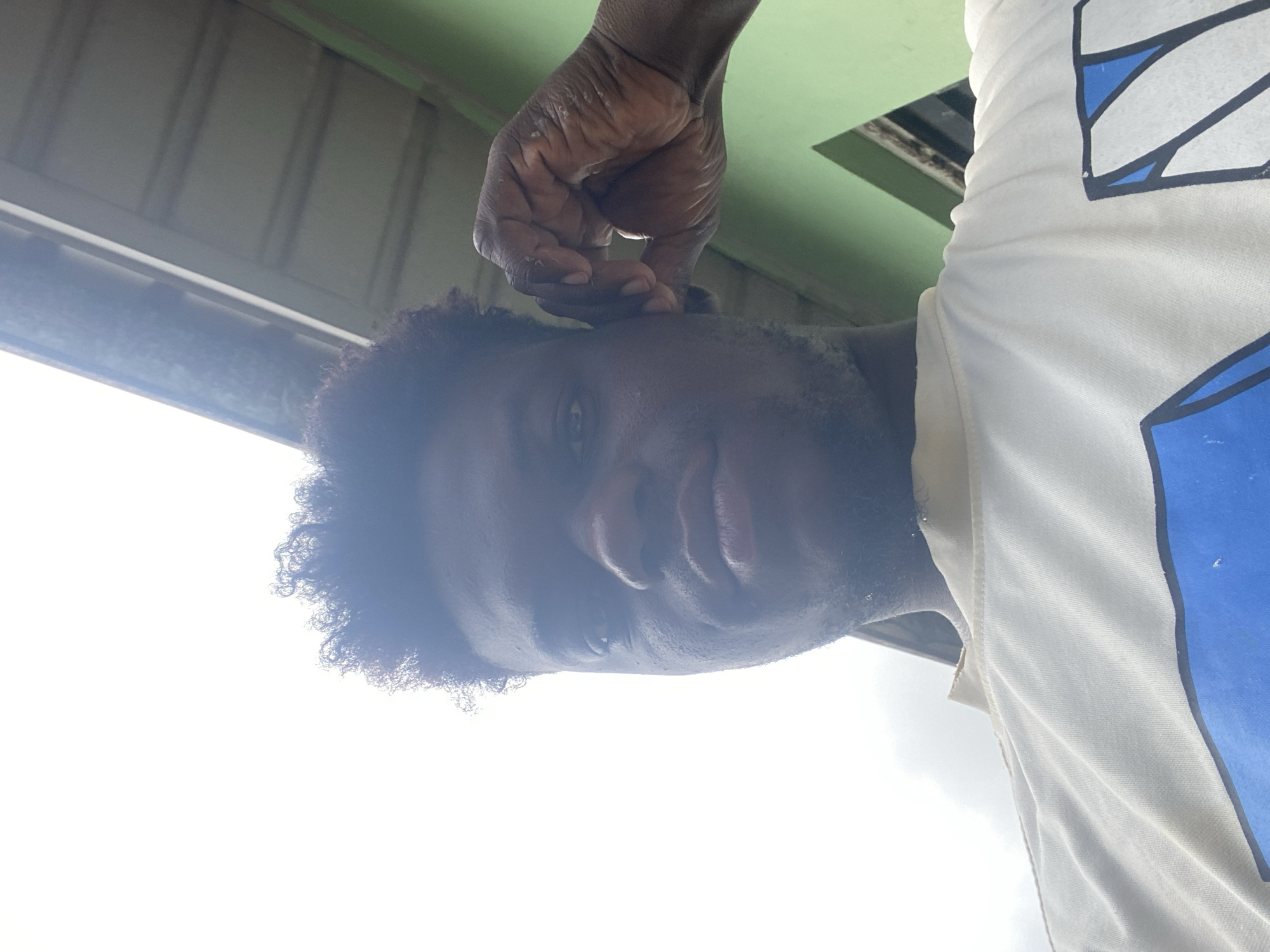 A man with dark skin and curly hair is depicted from a low angle, with a white shirt and blue accents visible. His hand is touching his face near his ear. The background includes a light green surface and a bright white sky. The location appears to be outdoors, possibly under a structure or awning, in an undefined urban setting in Unknown, Unknown.