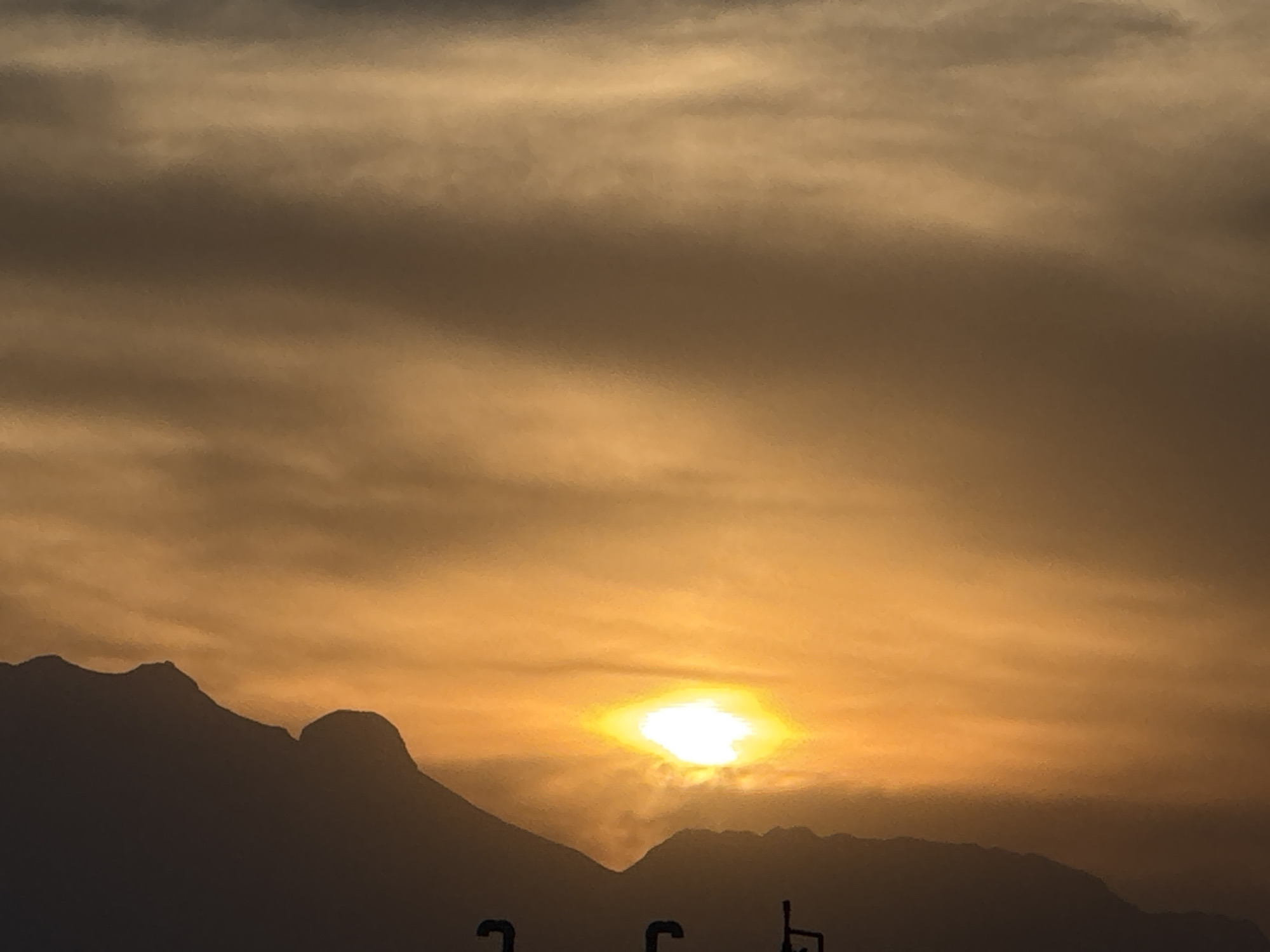 The image shows a sunrise or sunset over mountains near San Francisco Ocotlán, Mexico. The sky is filled with orange and brown clouds, and the sun is a bright white disc low on the horizon. The mountains are dark silhouettes against the bright sky. Some poles and wires can be seen in the very bottom part of the frame, partially obscuring the view.