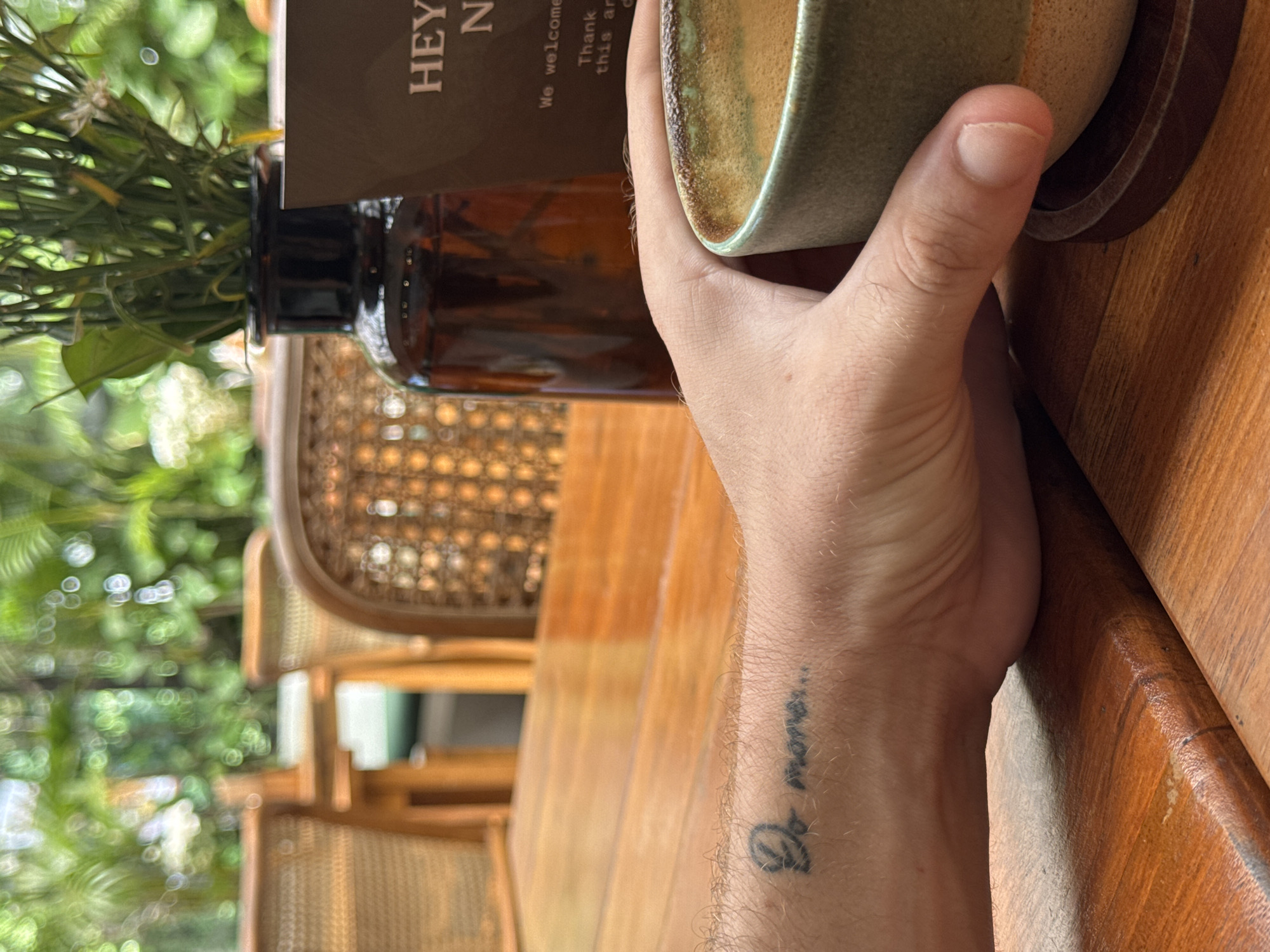 The scene appears to be a table setting at a cafe or restaurant, possibly located in Ungasan, Indonesia, given the tropical-like foliage visible through the blurred background. A fair-skinned hand holds a ceramic mug or cup with a brown base and green and brown interior. The forearm of the hand is visible, bearing a tattoo in black ink. The table is made of polished wood. To the left of the hand and cup is a brown glass bottle containing green foliage, which is partially obscured by a brown paper sign. The background features partially blurred woven-back chairs, and what looks like lush, green vegetation outside.
