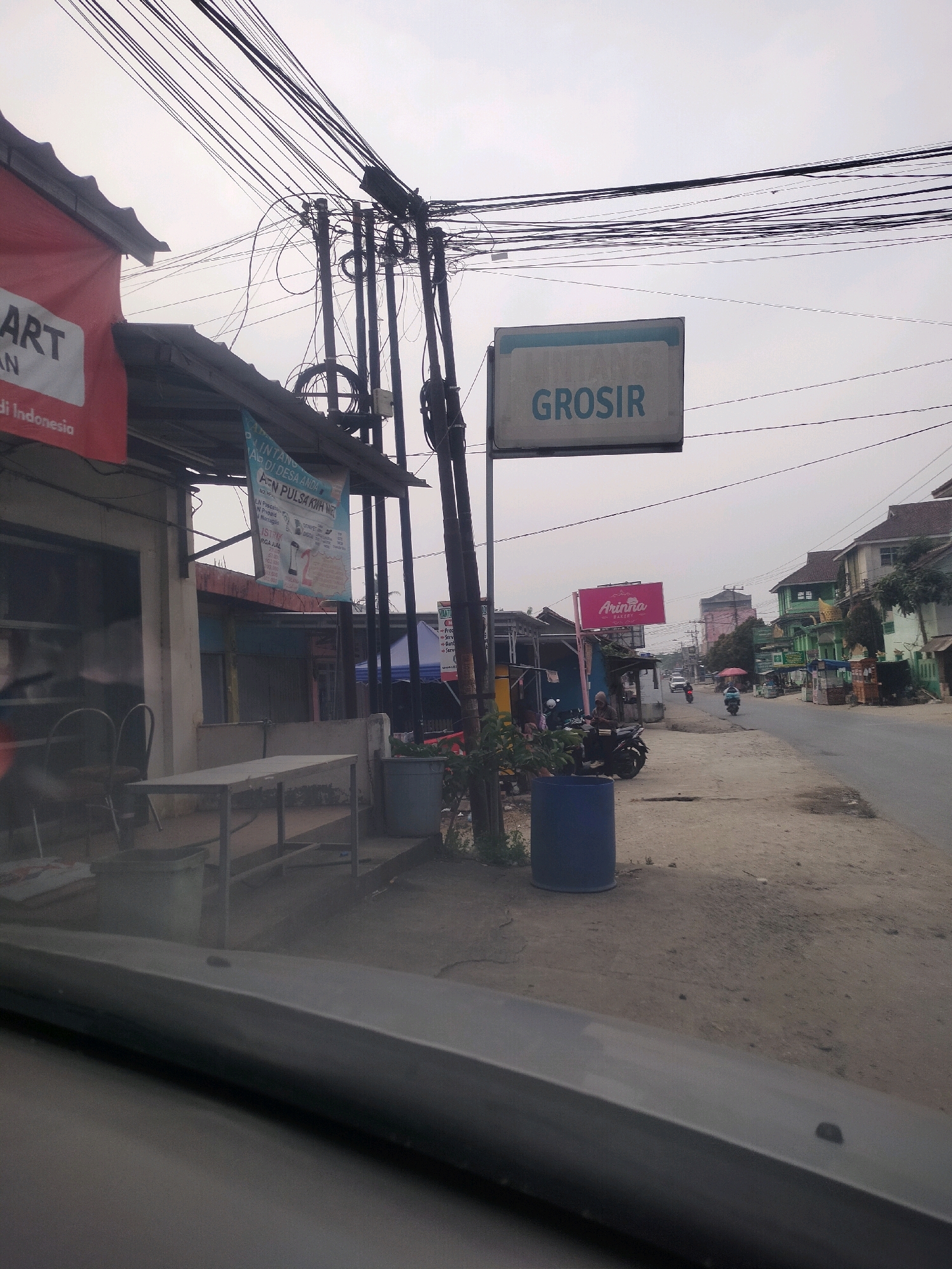 The image depicts a street scene in Wayakroy, Indonesia. In the foreground, a car's dashboard is visible. The street is lined with various buildings, including a store with a red awning and a sign that says "MART AN" and "di Indonesia". Another building has a blue awning and a banner advertising "PULSA MURAH". A prominent sign for "GROSIR" is visible, indicating a wholesale store. Further down the street, there are more shops and houses, some painted in green and yellow. Several motorcycles are parked along the roadside, and a few people are present, including a person sitting on a motorcycle. The sky is overcast, and electrical wires are strung across the poles, creating a cluttered overhead view. The ground appears to be a mix of dirt and paved road. There are also trash cans visible along the street. The overall impression is that of a busy, commercial street in a Southeast Asian town.