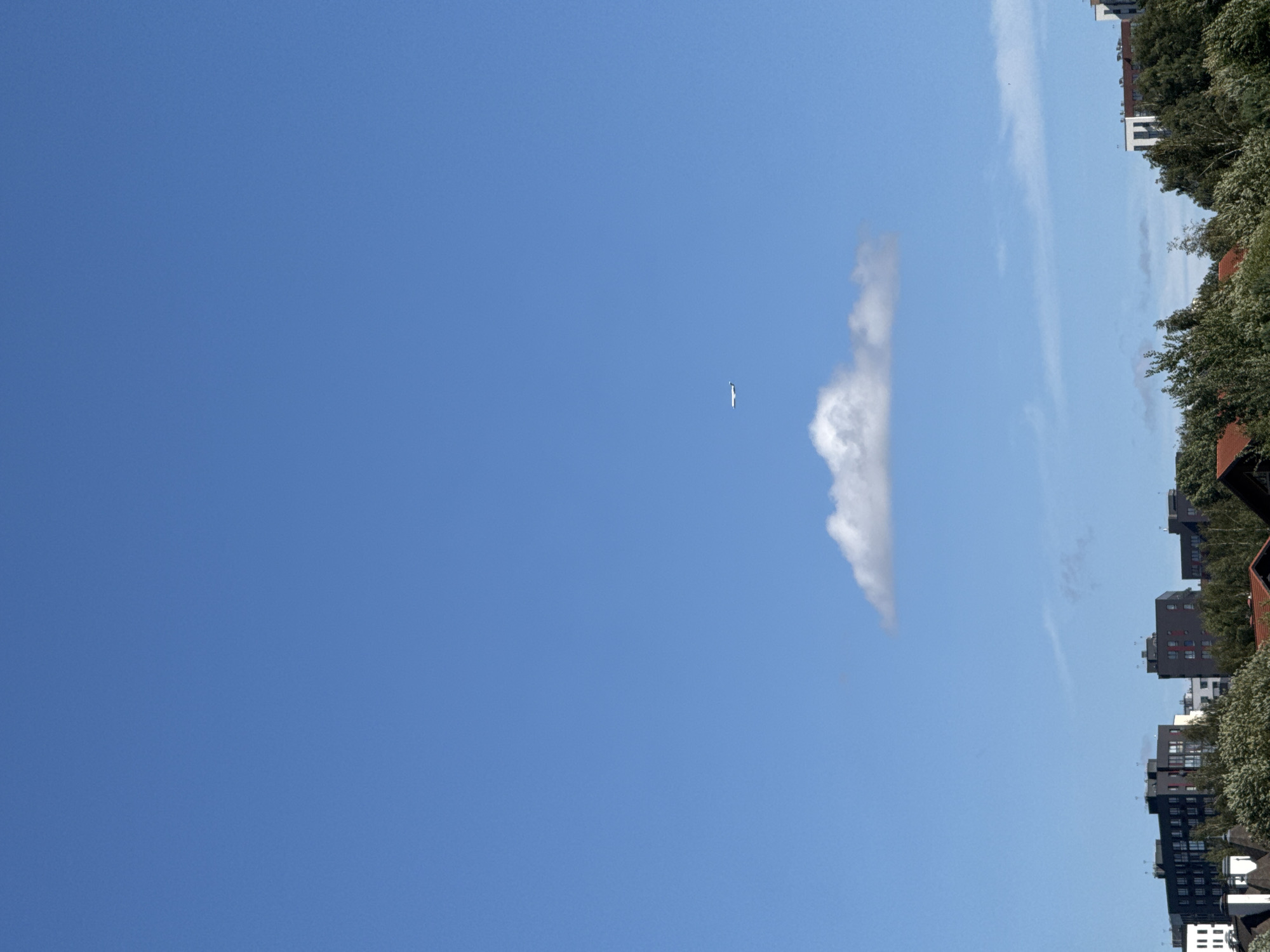 An image taken from a high vantage point overlooks a city skyline, likely in Kommunarka, Russia, under a clear blue sky. In the distance, a lone airplane is visible, flying against the expanse of the sky. Below the plane, a single, fluffy white cloud floats horizontally. The foreground is dominated by the roofs of buildings and lush green trees, suggesting a suburban or mixed-use urban environment.