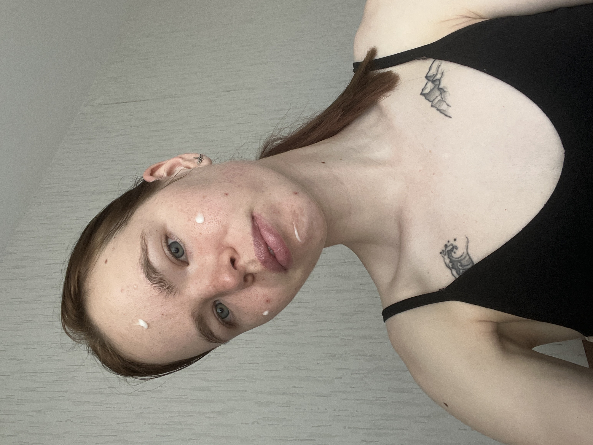 A young woman with fair skin and light blue eyes, wearing a black tank top, poses with a slight head tilt. White cream dots her forehead, cheek, and chin. Her brown hair is pulled back in a ponytail. Tattoos are visible on her upper chest and shoulder. The background is a textured, light-colored wall. There is no discernible evidence or indication that the image was taken in Perm, Russia.