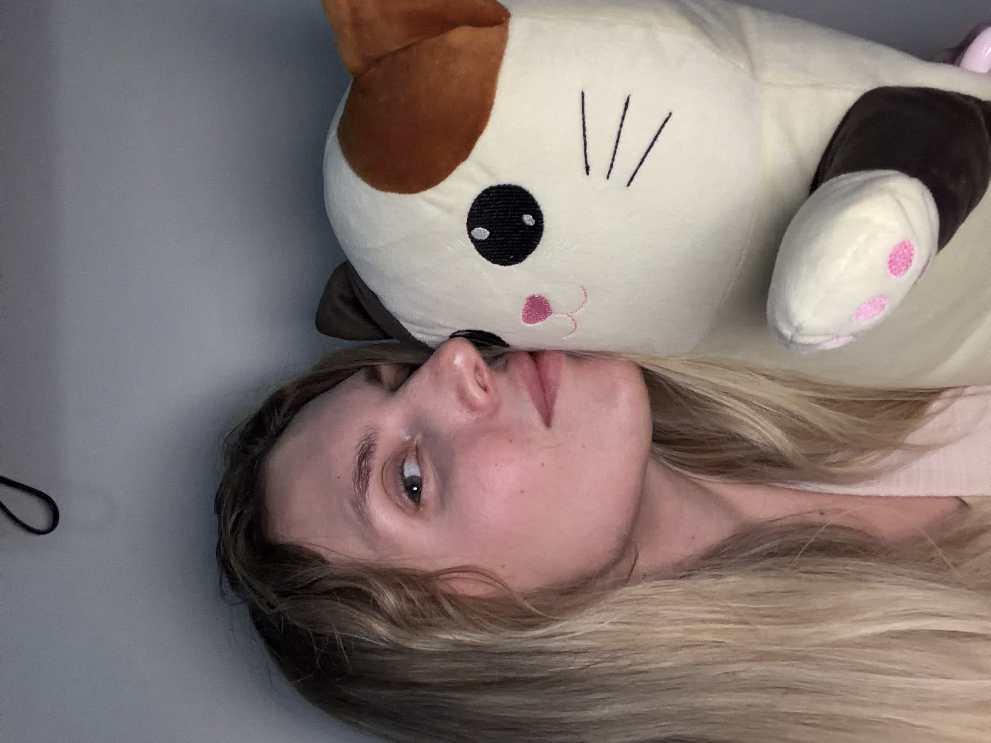 A close-up, rotated image shows a person resting alongside a large cat-shaped plush toy in an indoor setting. The person, a woman with long, blonde or light brown hair, is positioned on the left side of the frame, her head turned slightly to her left and looking upwards. Her visible eye is dark, and her expression appears calm and relaxed. Her fair skin is visible, and her hair flows out around her head.
Next to her, taking up a significant portion of the right side of the frame, is a cream-colored plush cat. This soft toy has prominent brown patches on its ears and across its back, and its visible front paw also has a brown section with pink embroidered paw pads. The cat's face features large, round black embroidered eyes with white highlights, a small pink embroidered nose, and a simple pink embroidered mouth. Three black embroidered lines representing whiskers are visible on its cheek.
The background is a plain, light gray surface, suggesting an intimate indoor environment like a bed or sofa. The lighting is soft and even, offering no specific cues about the time of day. There is no visible text. The overall scene conveys a feeling of comfort and relaxation, set in Budva, Montenegro.