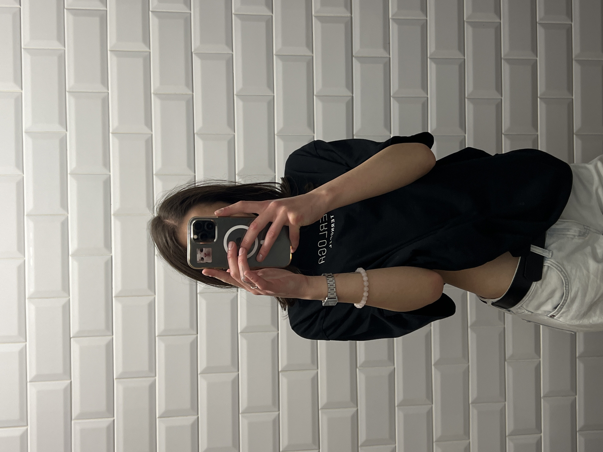 A young woman with brown hair is taking a mirror selfie in front of a white subway tile wall. She is wearing a black t-shirt and light-colored jeans with a black belt. On her left wrist, she has a silver watch and a pink beaded bracelet. She is holding a smartphone, which has a small sticker of a cat on the back. The image does not contain enough information to determine the location in Ternopil, Ukraine.