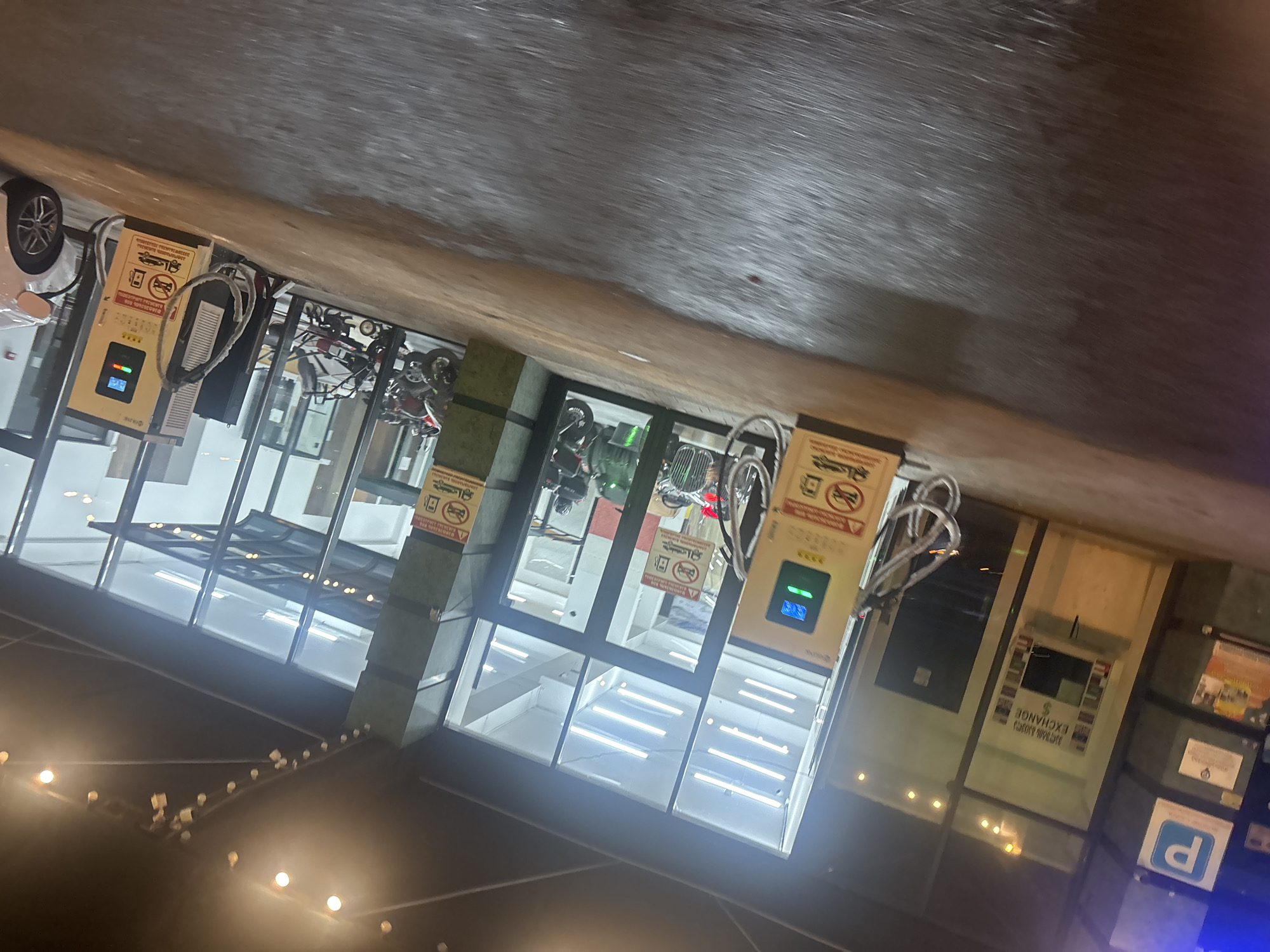 The image depicts a street scene in Tbilisi, Georgia. Three EV charging stations are lined up against a building with glass windows, each displaying operational information. A white car is parked next to one charging station. Several scooters and motorcycles are visible behind the windows of the building, possibly for sale or rent. The scene is illuminated by street lights and decorative lights strung overhead. A sign reading "Money Exchange" is affixed to the building. A blue and white parking sign is visible on the right.