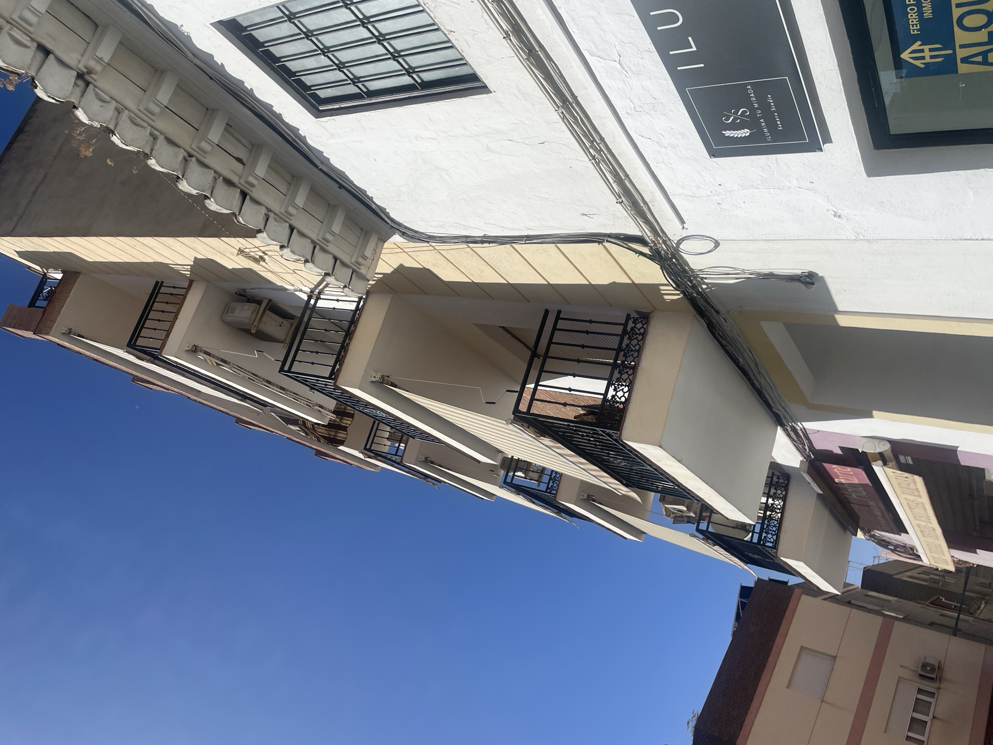 The image shows a street scene in Atarfe, Spain, under a clear blue sky. The perspective is looking up at buildings from a low angle. On the right is a white-painted building with a dark-framed window and a sign that reads "ILU MINA TU MIRADA." Further down the street, a portion of another building with a brown roof and light-colored facade is visible on the left. In the center, a multi-story building with balconies and awnings is depicted. The building has cream-colored walls and black metal railings on its balconies. Electrical wires are visible running along the facade of the white building. A sign for "FERRO REAL ESTATE ALQU" is partially visible in the lower right corner, suggesting a commercial area. No people are visible in the image.