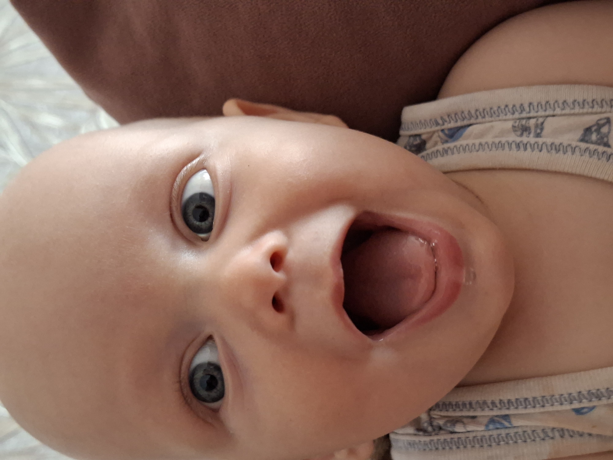 A close-up, eye-level shot shows a baby's face with blue eyes, an open mouth, and a bald head. The baby is wearing a white top with blue and black patterns. The background is blurred, with a brown surface on the right and a white, textured surface on the left. There is a slight sheen on the baby's chin and lip, suggesting moisture or saliva.