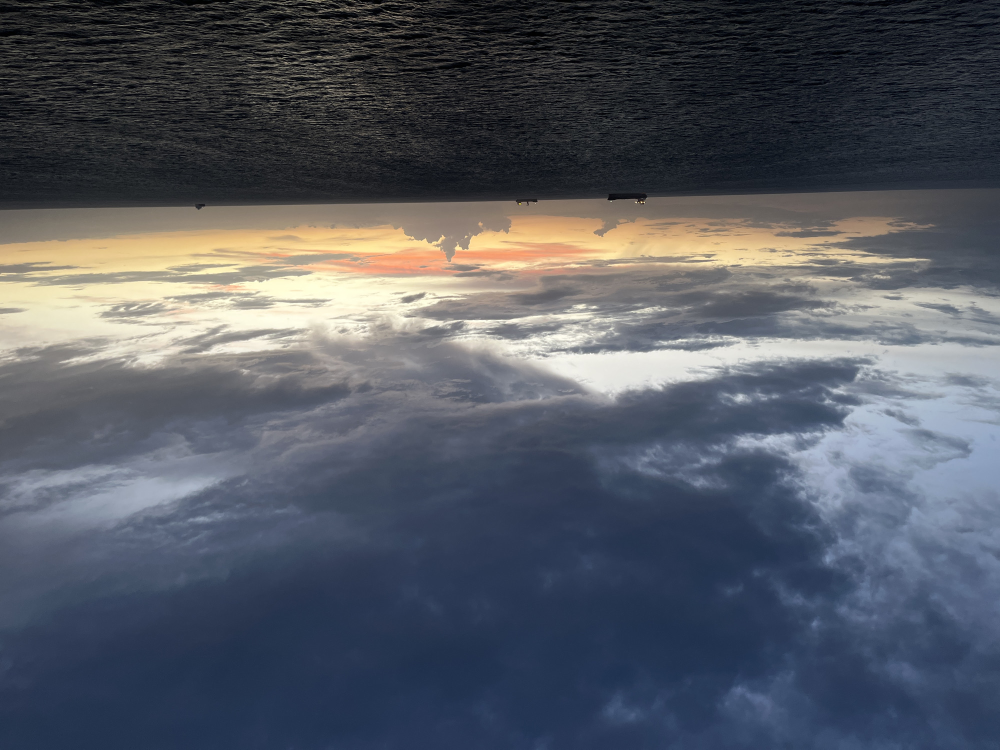 The image presents an upside-down view of a seascape at twilight. The bottom half displays a cloudy sky with varying shades of gray and blue, darkening towards the "lower" edge. A layer of orange and yellow hues marks the horizon line, suggesting the setting or rising sun. Above the horizon, the upper half of the image depicts a dark, rippled body of water, possibly the ocean. Three silhouettes resembling boats or ships are visible on the horizon line. The location is unknown.
