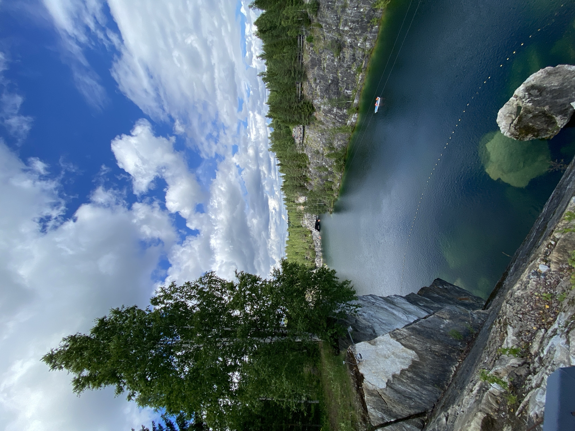 This image depicts a scenic view of the Ruskeala Mountain Park, a former marble quarry in Ruskeala, Russia. The foreground is dominated by a steep, rocky cliff face with visible layers of white and grey marble. A large birch tree with white bark and green foliage grows precariously from the edge of the cliff, its branches extending over the water.
The middle ground features a serene, deep blue lake, likely a flooded part of the quarry. The water is clear, with visible underwater features and a few yellow buoys forming a line across the surface. A small white boat with red trim is visible on the lake. The surrounding landscape is characterized by rugged, grey rock formations that form the steep banks of the quarry.
In the background, a dense forest of green coniferous and deciduous trees covers the hills. A narrow walkway or path is visible along the top of the quarry wall, with some structures or viewing platforms visible. Further in the distance, a tunnel entrance can be seen carved into the rock face, hinting at the historical use of the quarry for marble extraction. The sky above is a vibrant blue, dotted with fluffy white clouds, suggesting a clear and pleasant day. The overall impression is one of natural beauty and historical significance, showcasing the transformation of an industrial site into a popular tourist destination.