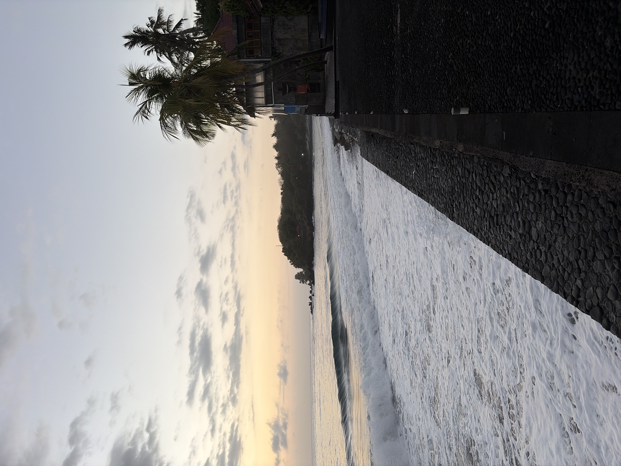 The image depicts a coastal scene at sunrise in Kubu, Indonesia. The sky is a soft gradient of pale blue and warm yellow, with scattered clouds. Gentle waves with white foam are rolling onto a dark, pebbled shoreline. To the right of the frame, a building with a red-tiled roof and dark walls stands next to two palm trees. A dark, paved walkway lines the edge of the property, separating it from the beach. The overall atmosphere is tranquil and serene, characteristic of a tropical morning.