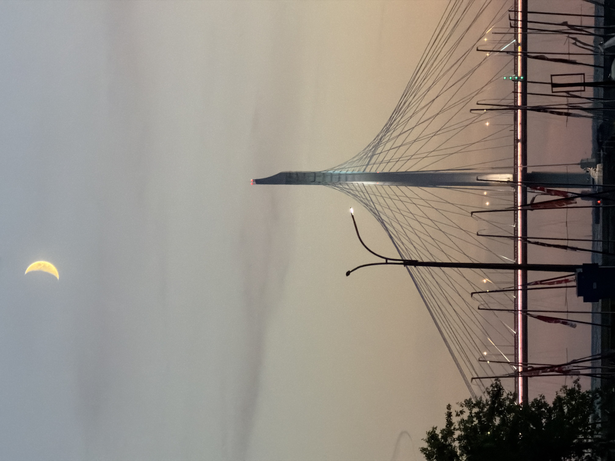 A crescent moon is visible in the pale sky above the Western High-Speed Diameter bridge in Saint Petersburg, Russia. The bridge's distinctive pylon, illuminated by red lights at its apex, dominates the center of the frame. The bridge's suspension cables fan out from the pylon, crisscrossing the sky. In the foreground, a lamppost stands to the left of the bridge, and silhouetted flags on poles are clustered near the bottom of the image. The sky is a gradient of soft grays and pinks, indicating either dawn or dusk.