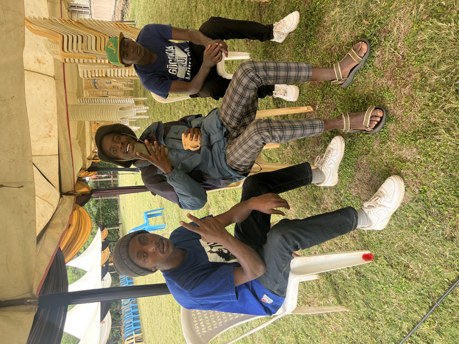 The image, taken in Makuyu, Kenya, shows three young men sitting on white plastic chairs outdoors under a large tent during daytime. The ground is covered with grass, and stacks of similar chairs are visible in the background, suggesting an event setting.
From top to bottom in the upright image orientation:
The man at the top wears a blue t-shirt with the text "GÜCKNA RUI MUMBA" printed across the front, black pants, and a green and yellow baseball cap. He appears to be resting with his eyes closed, wearing white sneakers.
The man in the middle wears a blue jacket over a t-shirt, distinctive plaid trousers, and a dark bucket hat. He holds a smartphone in his left hand and a yellow mug in his right, smiling brightly at the camera. Partial text "GUCCIA RUI..." is visible on his t-shirt. He wears beige and black sandals.
The man at the bottom wears a blue t-shirt, dark jeans, and a dark beanie. He is also smiling at the camera and making a peace sign with his right hand. He wears white sneakers.
The scene conveys a relaxed and casual atmosphere under natural daylight.