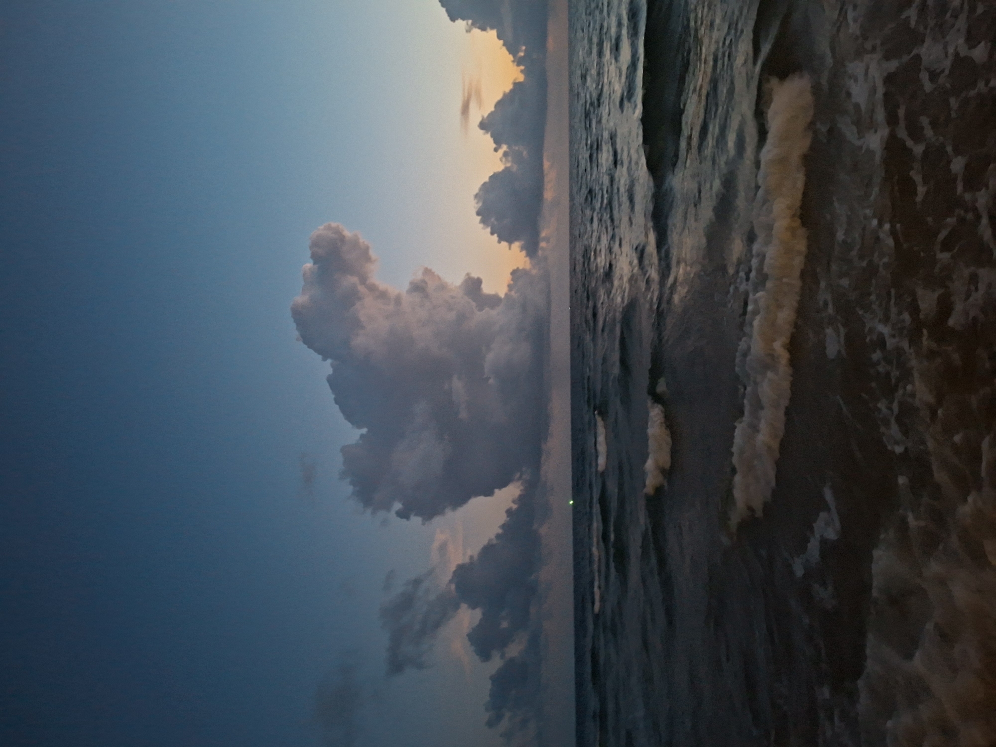 The image shows a body of water with waves, likely the ocean, under a twilight sky. Several clouds are visible, with one large, fluffy cloud dominating the upper center of the frame. There is a faint orange glow on the horizon, suggesting either sunrise or sunset. A small green light is visible on the water in the distance. The overall scene is calm and serene. There are no people or specific structures identifiable in the image. The location is described as Unknown, Unknown.
