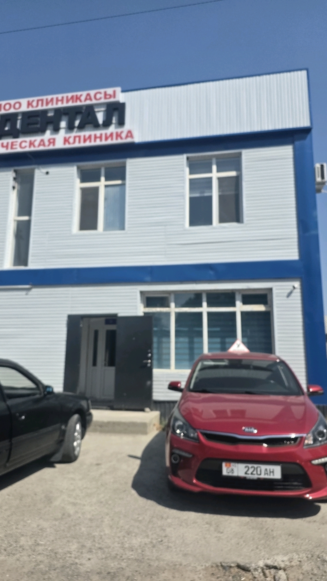The image shows the facade of a two-story building with a blue stripe running horizontally across the upper part of the first floor. Above this stripe, on the second story, is a white section with lettering. The visible lettering, in dark gray and red, reads "ДЕНТАЛ" in large dark gray letters and "ЧЕСКАЯ КЛИНИКА" in red letters below it. Above "ДЕНТАЛ" is "ОО КЛИНИКАСЫ". This suggests the building houses a dental clinic. The building has several windows on both floors. On the first floor, there is an open doorway.
In front of the building, two cars are parked. On the left, a black car is partially visible. On the right, a red car is parked closer to the building. The license plate of the red car is visible and reads "08 220 AH". A small red triangular sign is visible on the roof of the red car. The ground in front of the building appears to be asphalt or a similar surface. The sky is clear and blue. The overall impression is an exterior shot of a commercial establishment in what appears to be an urban or suburban setting.