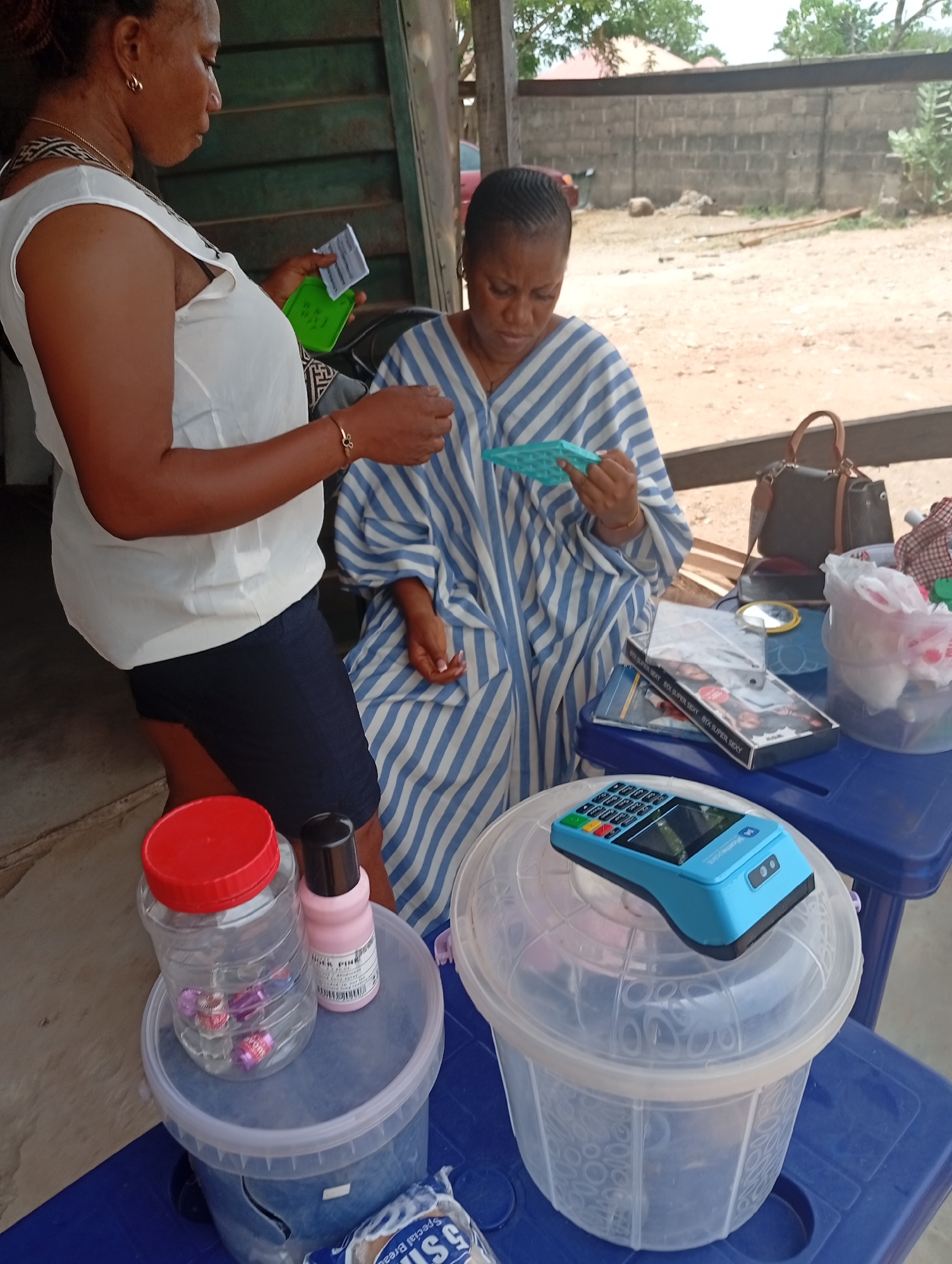 Two women are engaged in a transaction at an outdoor stall in what is identified as Adankolo, Nigeria. The woman on the left is standing, wearing a white top and dark skirt, holding papers in her hand. She is interacting with the seated woman, who is wearing a blue and white striped garment and holding a small blue item. A table in front of the seated woman displays items for sale, including cosmetics, a mirror, and packaged goods. A blue credit card terminal rests on top of a plastic container. The stall appears to be set up in an outdoor, semi-shaded area. A red-lidded jar and a pink bottle sit on a smaller container on a blue table. Bags and personal items are also visible on the table. The background shows an outdoor area with a building visible in the distance.