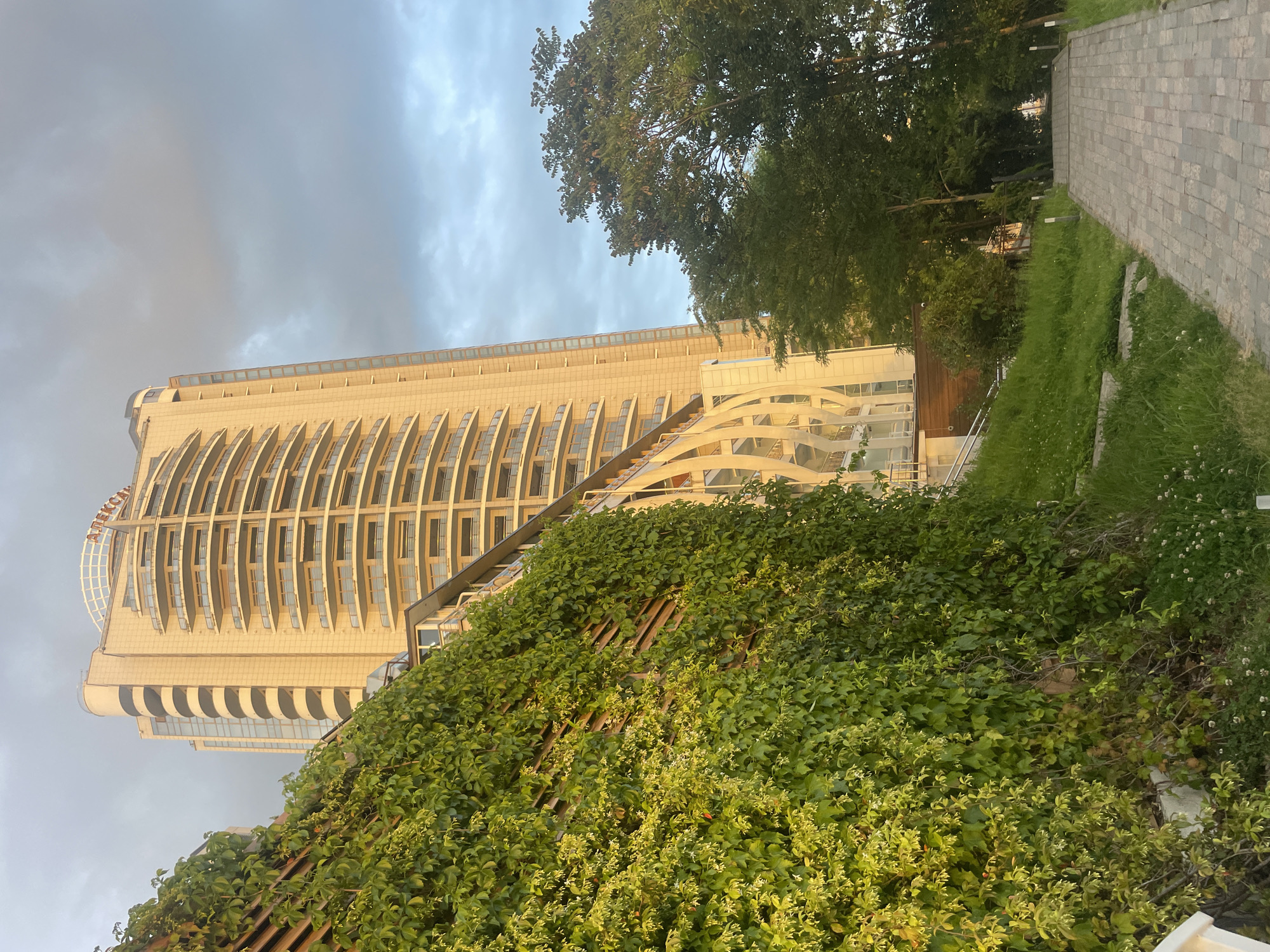 A slightly tilted eye-level shot showcases the Aleksandriyskiy Mayak (Александрийский маяк) building in Sochi, Russia, towering behind a wall covered in lush green vegetation. The building's architectural design features a curved facade with multiple floors and a glass dome on top. To the right of the building, trees are visible, and a paved walkway extends into the distance. Overcast skies provide a soft light to the scene. The dense greenery in the foreground contrasts with the building's facade, creating a visual interplay of nature and architecture.
